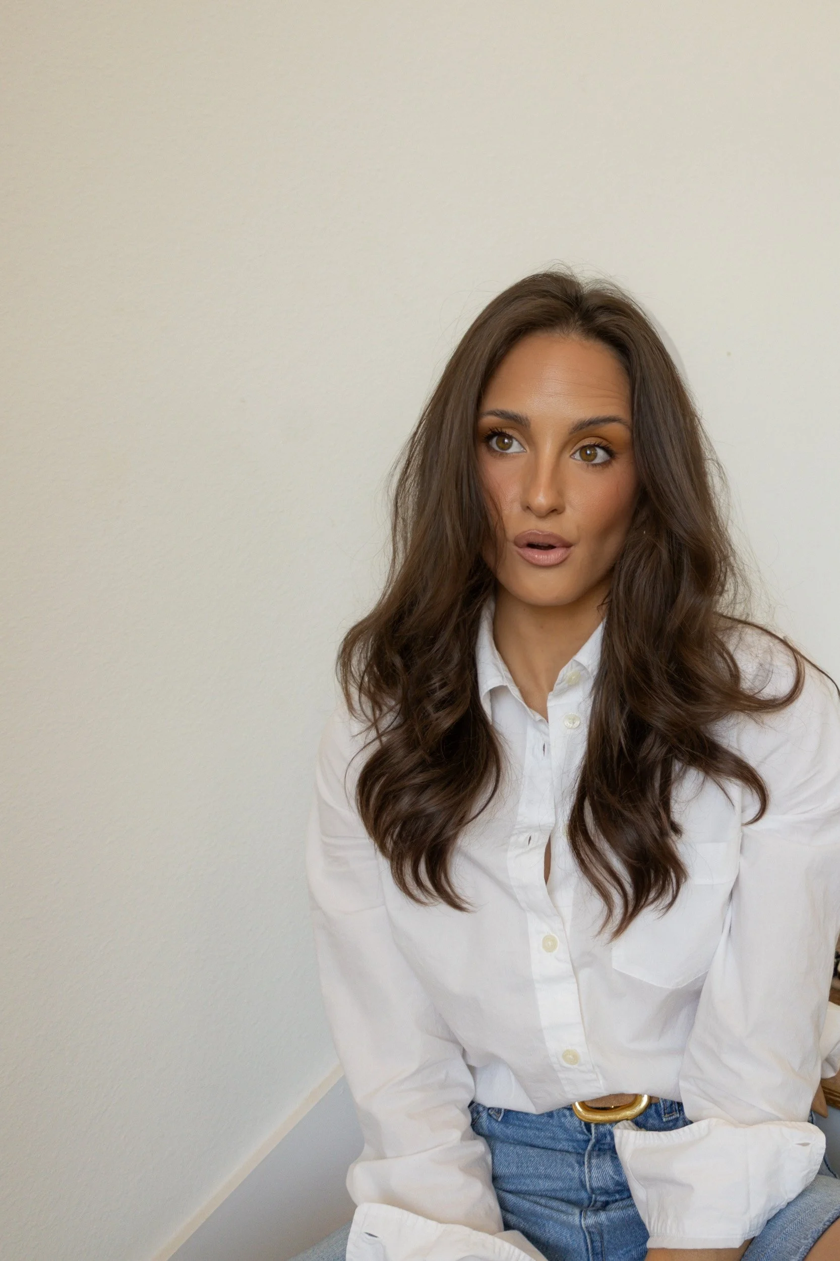 A woman with long wavy brown hair wearing a white button-up shirt and blue jeans sitting in front of a plain off-white wall.