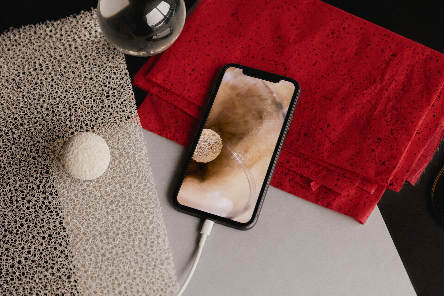 A smartphone connected to a charger resting on a surface beside a textured decorative object and a spherical object, with a stack of red napkins or paper towels nearby.