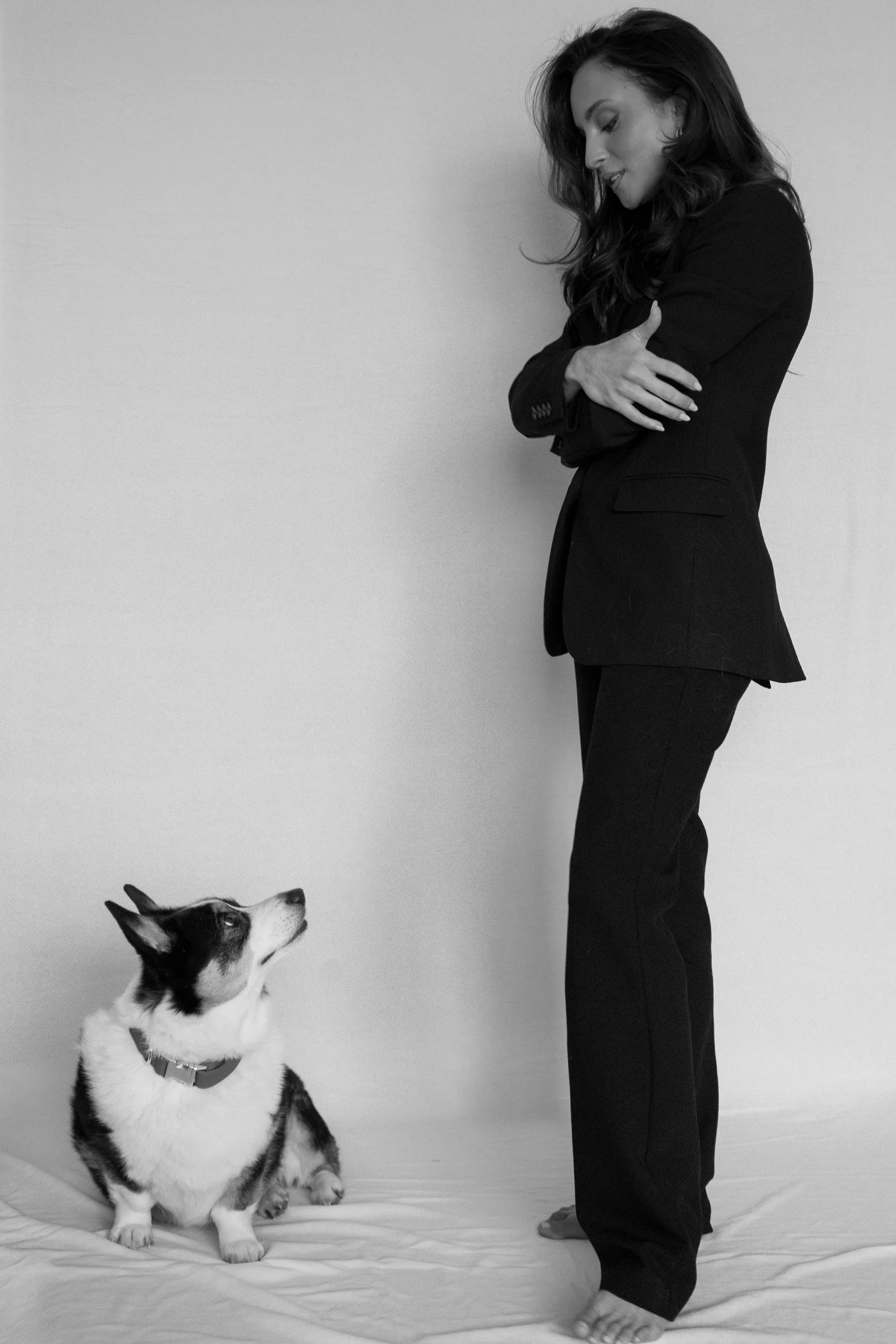 A woman with long dark hair wearing a black blazer and black pants standing next to a sitting husky dog, both on a plain floor, against a plain wall background.