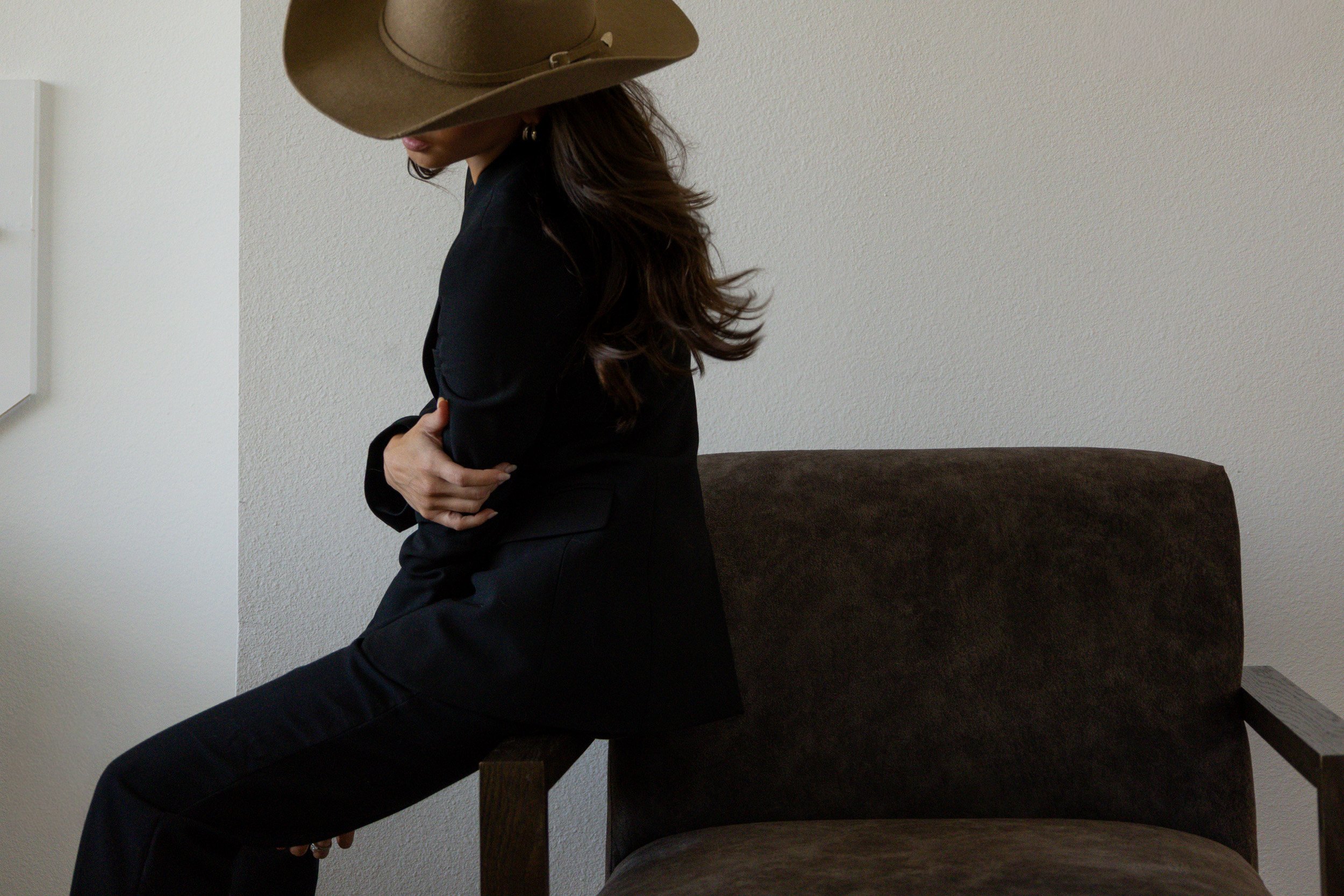 A woman wearing a large tan cowboy hat and black blazer, sitting on a brown upholstered chair against a beige wall, with her arms crossed and head slightly tilted down.
