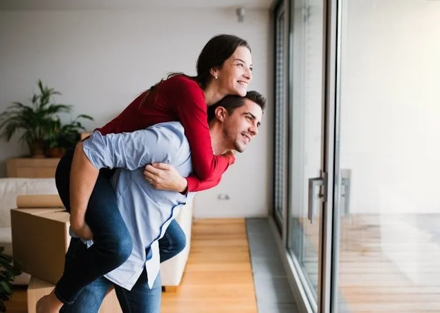 A happy couple enjoying a moment together indoors, with the woman on the man's back looking out a large window.