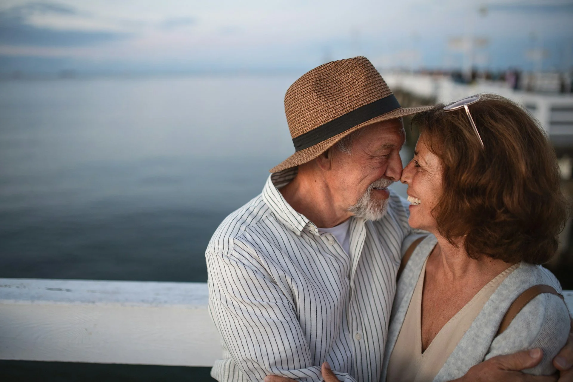 An elderly couple is smiling and touching foreheads near a body of water, with boats and a dock in the background.