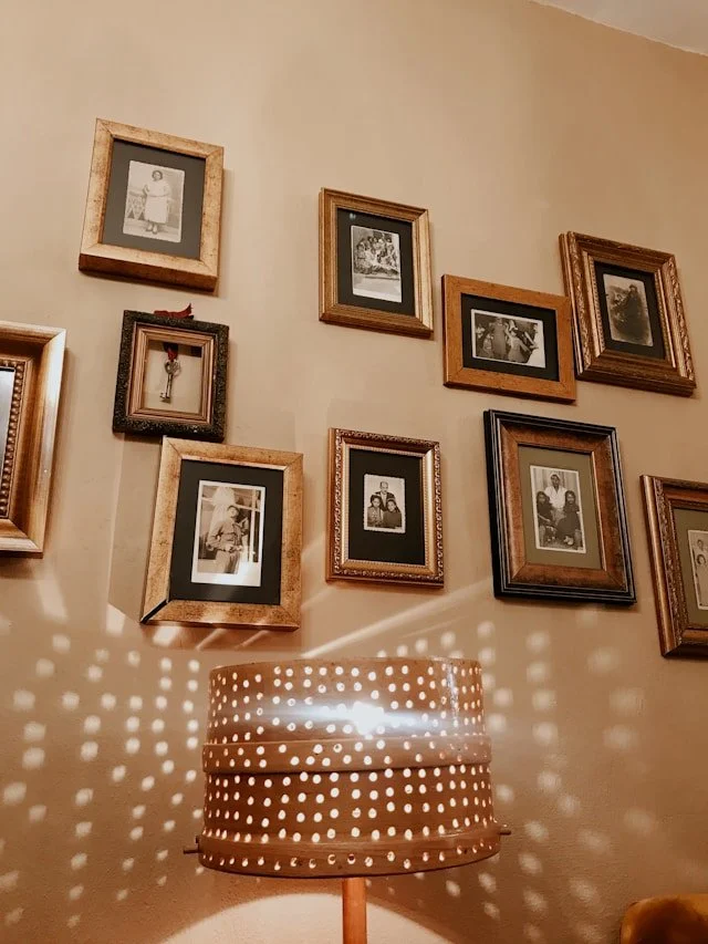 A wall decorated with various framed black-and-white photographs arranged in a gallery-style display. Below the photos is a lamp with a perforated shade, casting dotted light patterns on the wall.