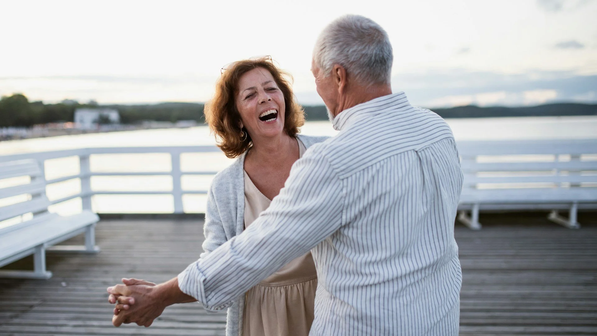 An elderly couple dancing and laughing on a wooden dock by the water during the day, with benches and a distant shoreline.