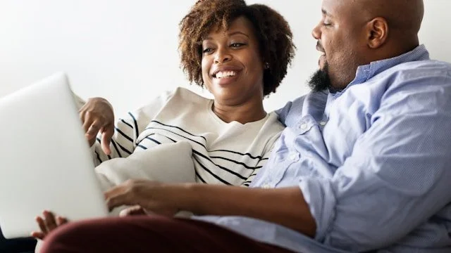 A smiling couple sitting close together, looking at a laptop.