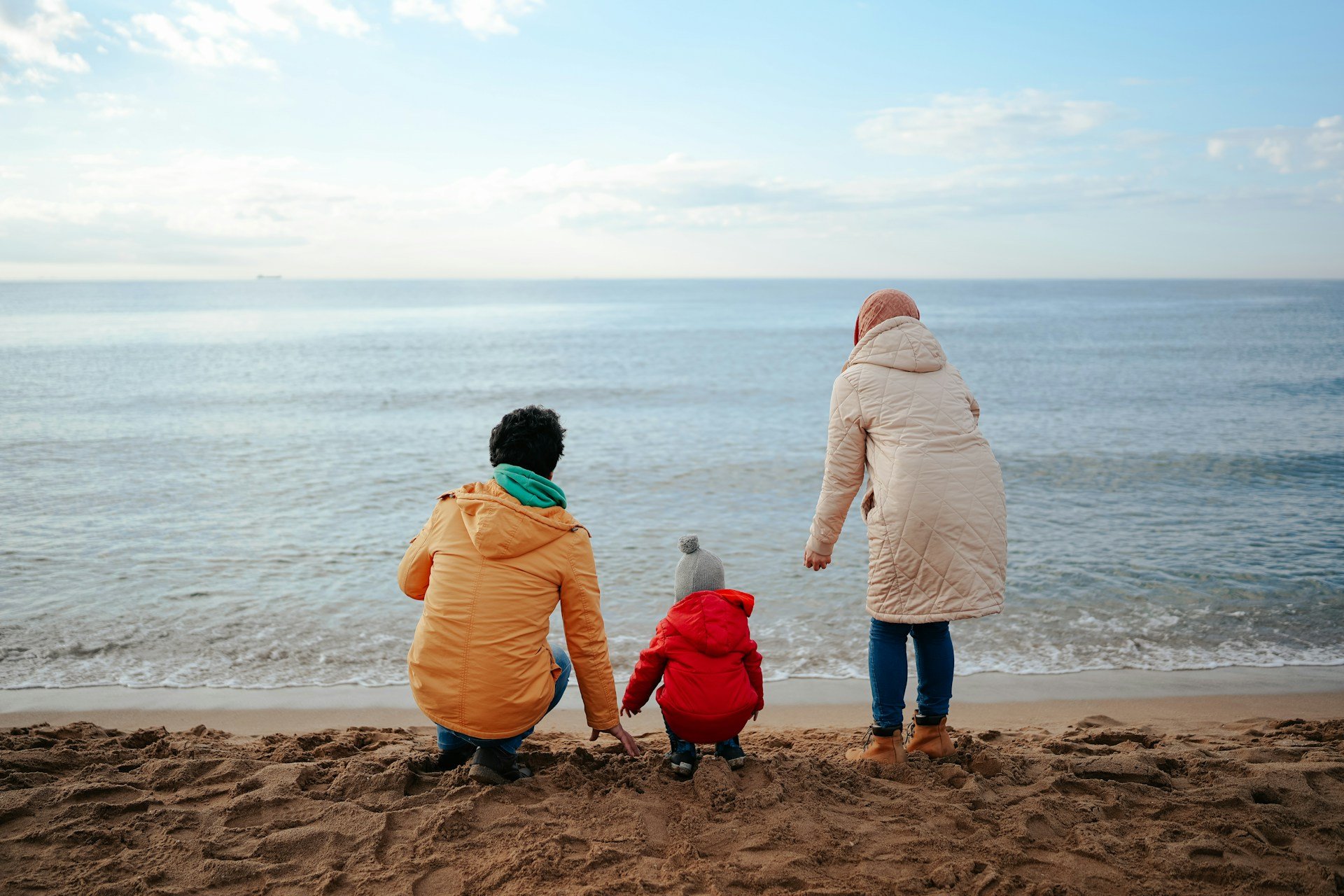 Four people dressed in winter clothing on a sandy beach, sitting and standing by the water, with a calm sea and a partially cloudy sky in the background.