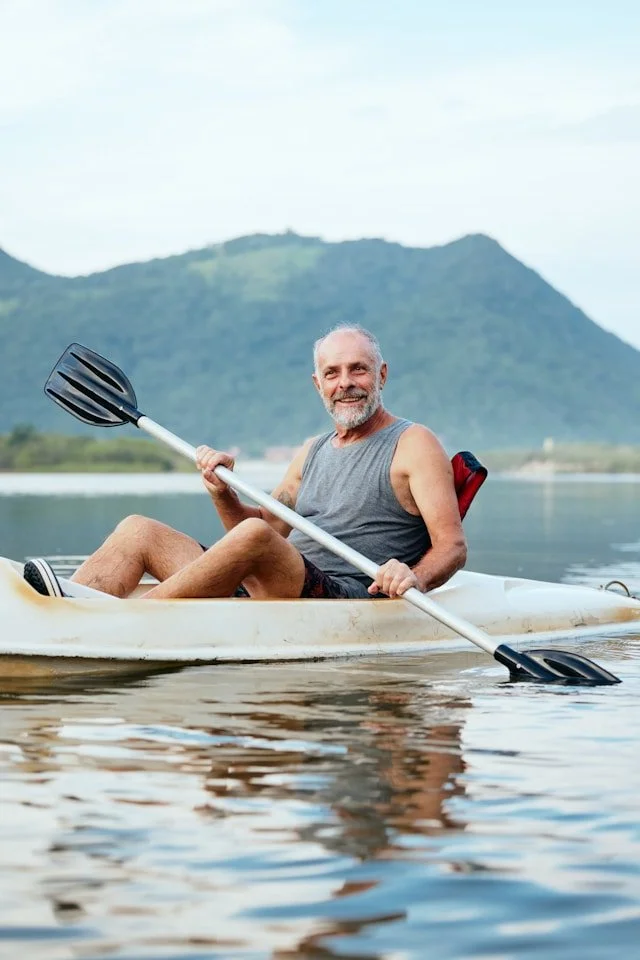 A smiling man with gray hair and a beard kayaking on a lake with a mountain in the background.