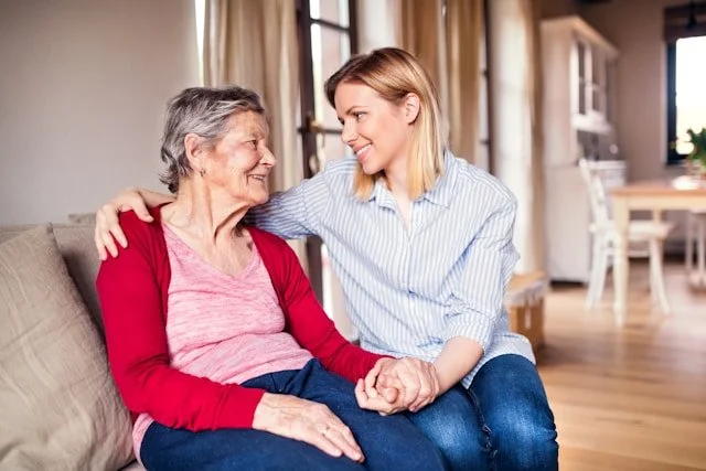 A young woman and an elderly woman sitting on a sofa, smiling and holding hands, in a cozy living room with natural light.