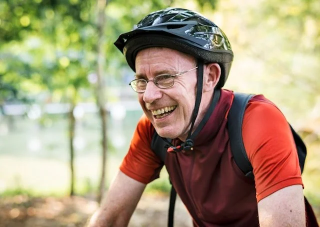 A man wearing glasses, a helmet, and a red cycling jersey smiling outdoors in a park with trees in the background.