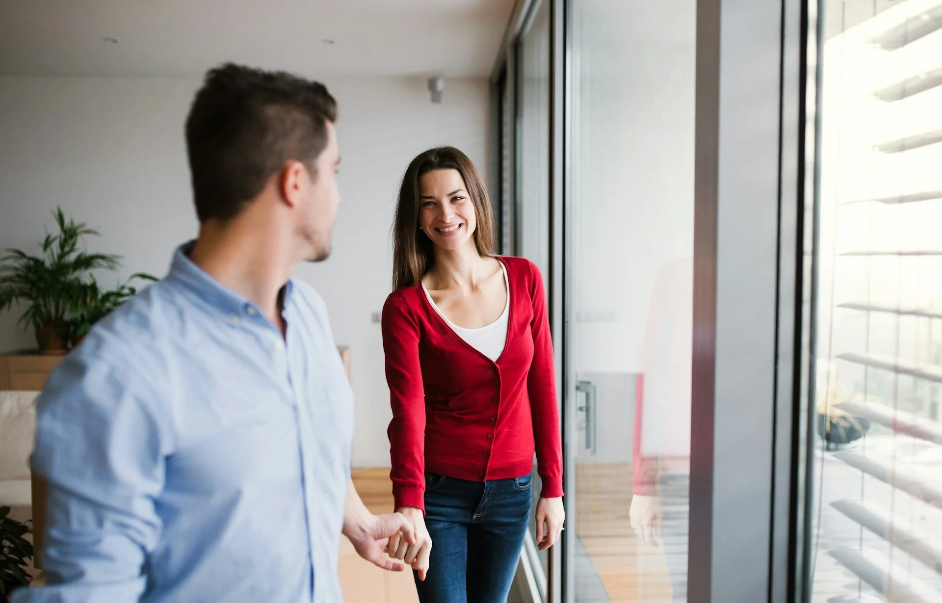 A man and woman smiling and holding hands near a large glass window in a bright modern room.