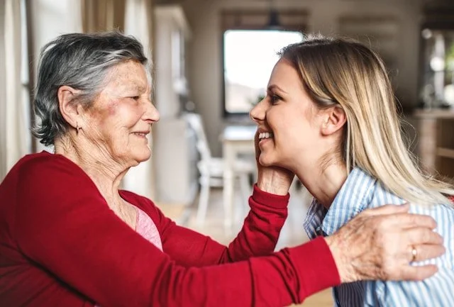 A young woman and an elderly woman smiling and engaging in an affectionate moment indoors.