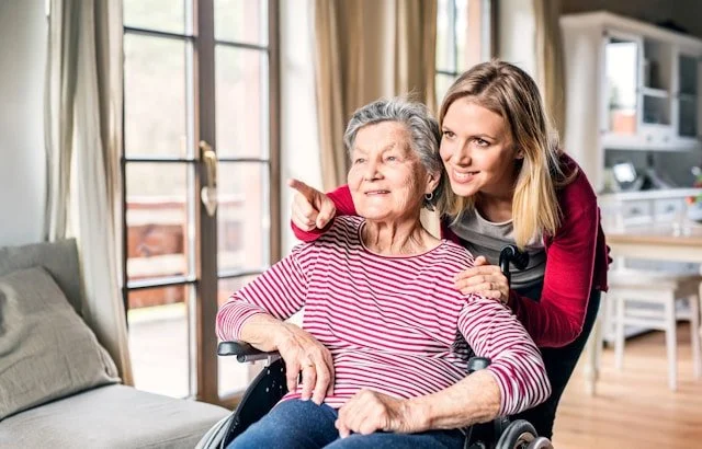 A young woman pushes an elderly woman in a wheelchair inside a bright living room.