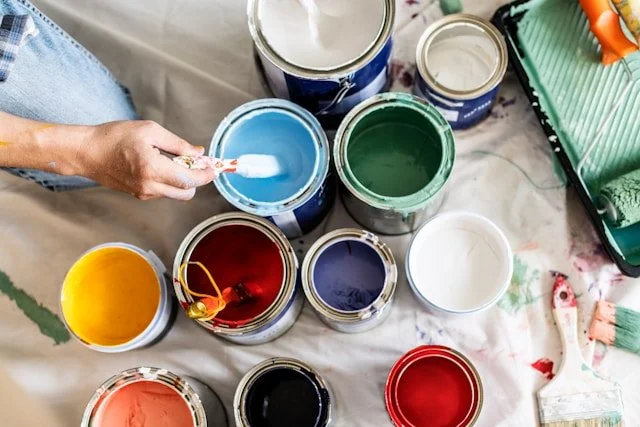 Person holding a paintbrush above open cans of blue, green, yellow, red, black, white, and coral paint on a table covered with a cloth with paint splatters.