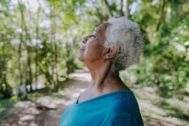 An elderly woman with gray curly hair standing outdoors on a forest trail, smiling with eyes closed, enjoying the sunshine.