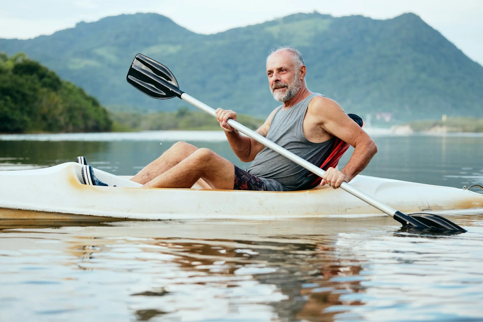 An elderly man with a beard kayaking on a calm river with mountains and greenery in the background.