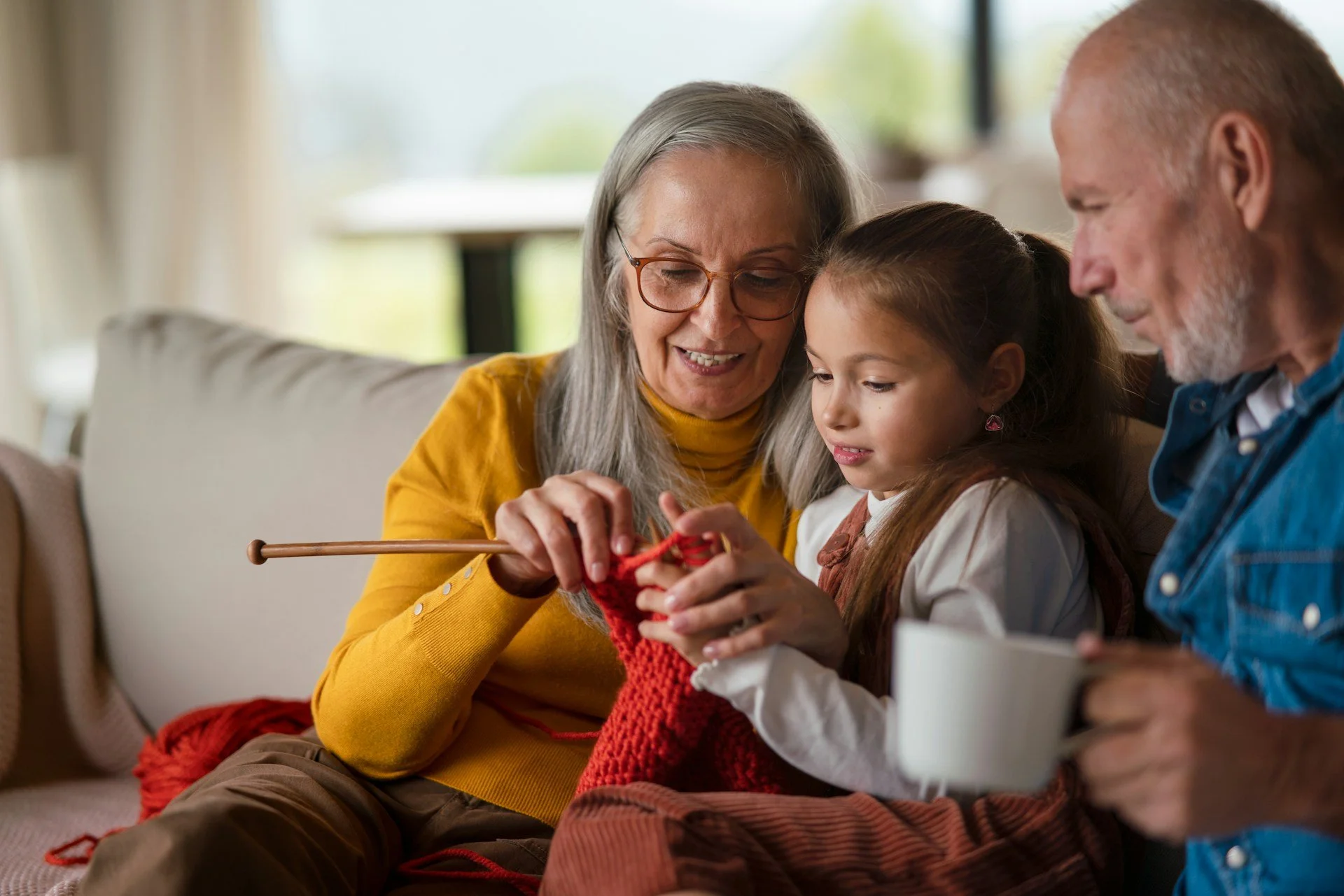 Grandparents and a young girl knitting together on a couch in a bright living room.
