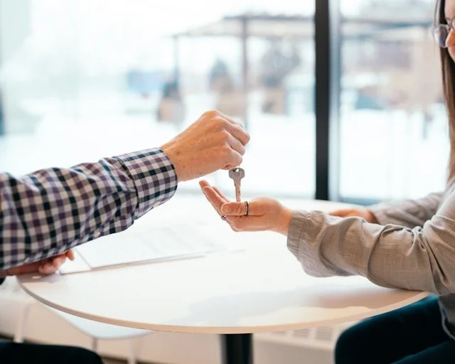 Person handing over car keys to another person indoors with large windows in the background.