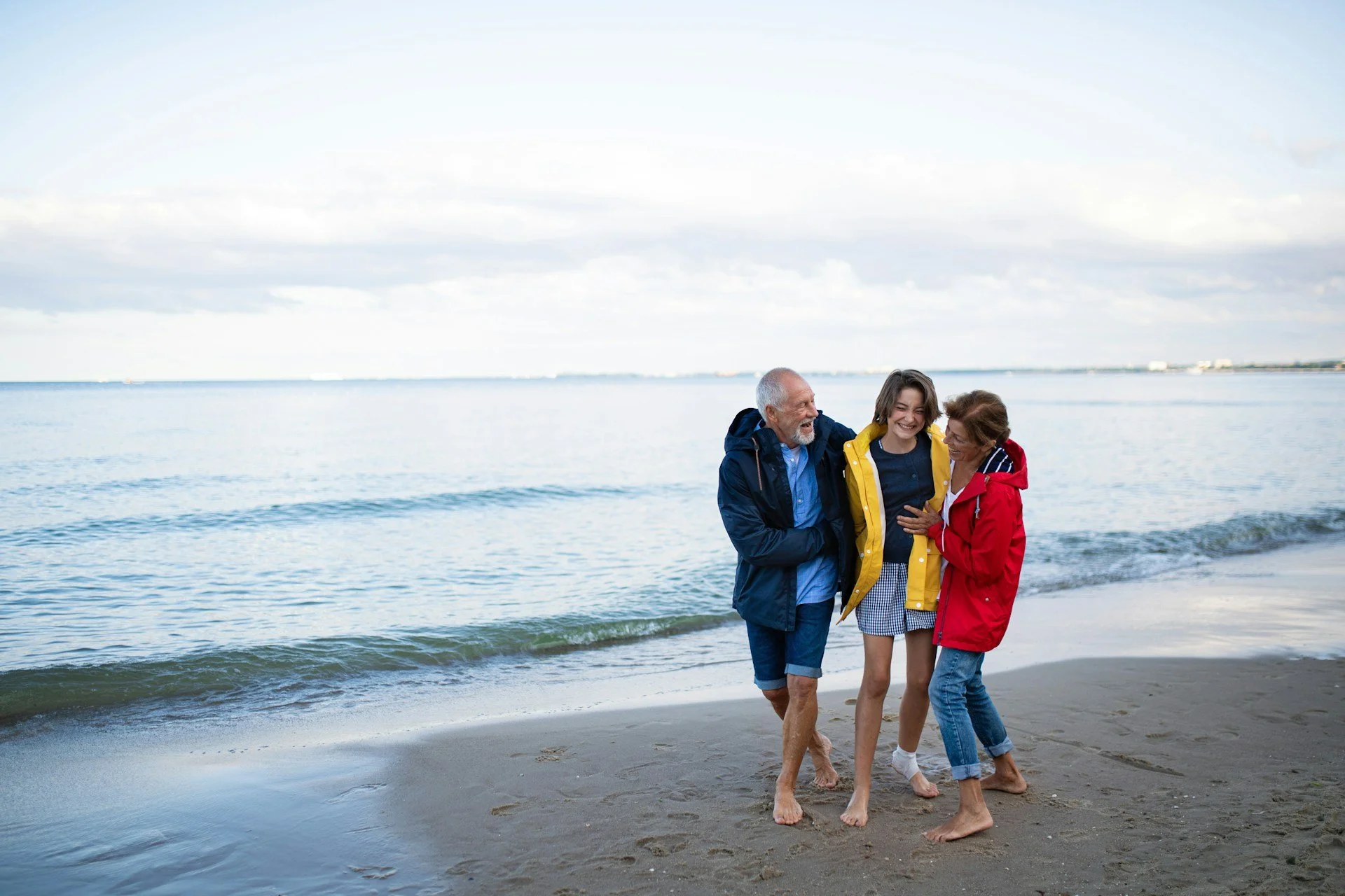 Three people, two women and a man, walking barefoot on the beach near the water, smiling, with an overcast sky in the background.