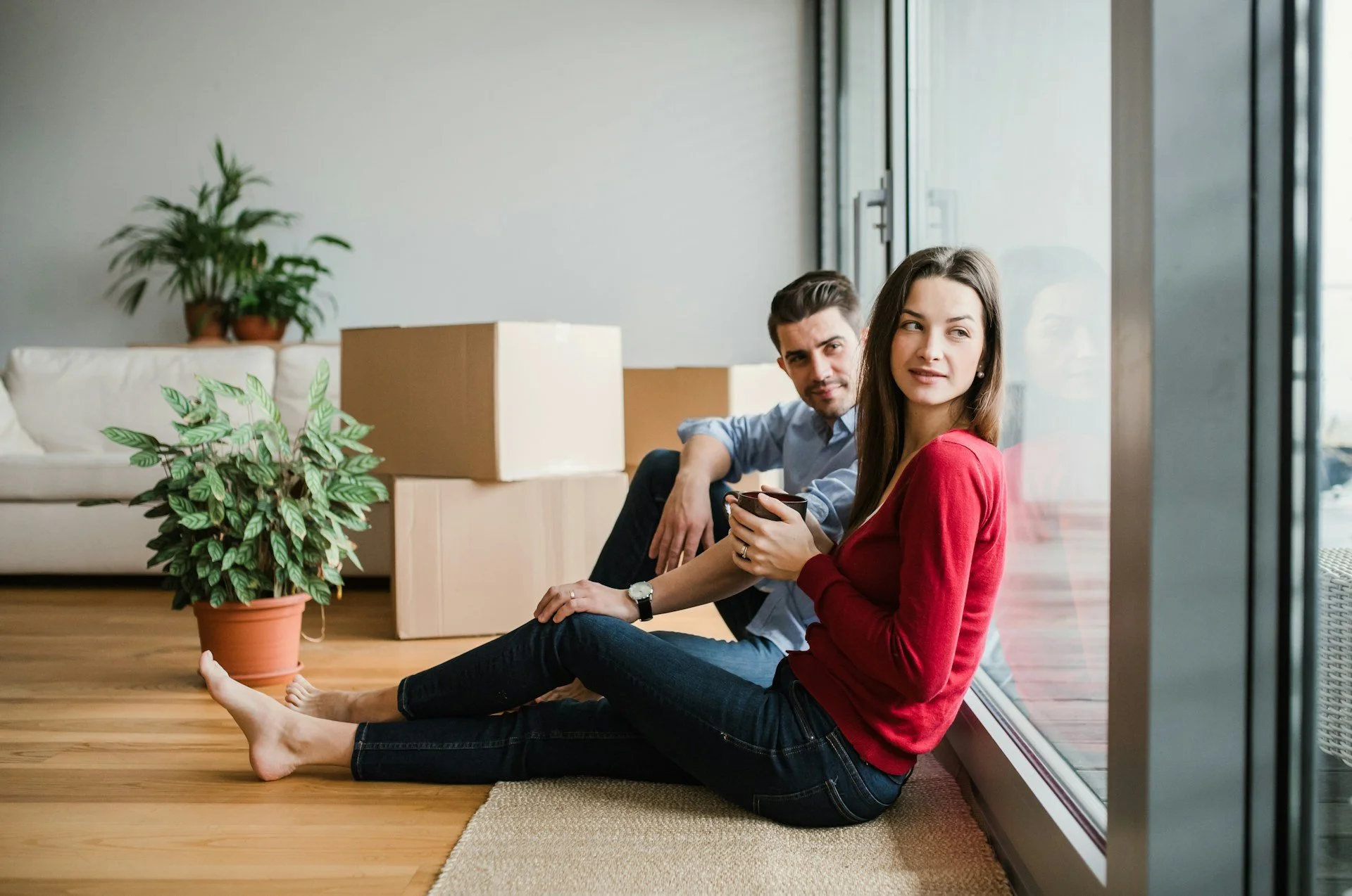 A man and woman sitting on the wooden floor near a glass wall, with cardboard boxes and potted plants in the background, suggesting they are in a new apartment or house.
