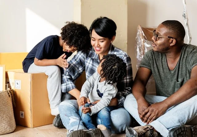 A happy multigenerational family sitting on the floor of a new home. A woman holds a young girl, a boy leans on a cardboard box, and a man sits beside them, all smiling.