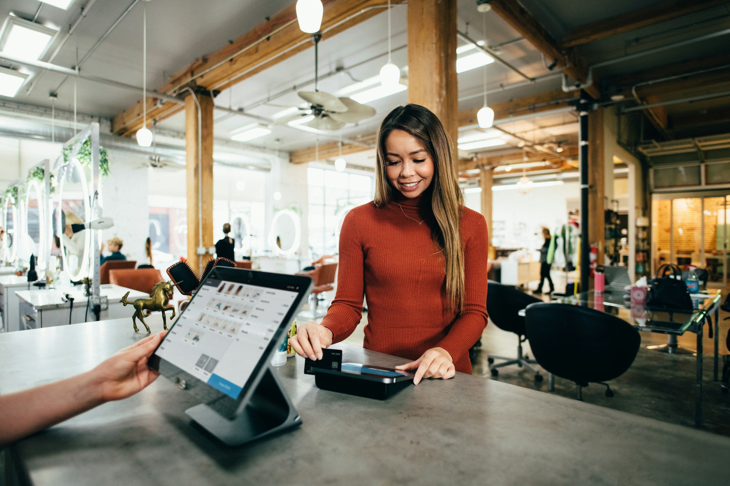 A woman in a red long-sleeve shirt is smiling and paying at a cashier in a modern interior space with wooden beams and large windows; a person holds a tablet.