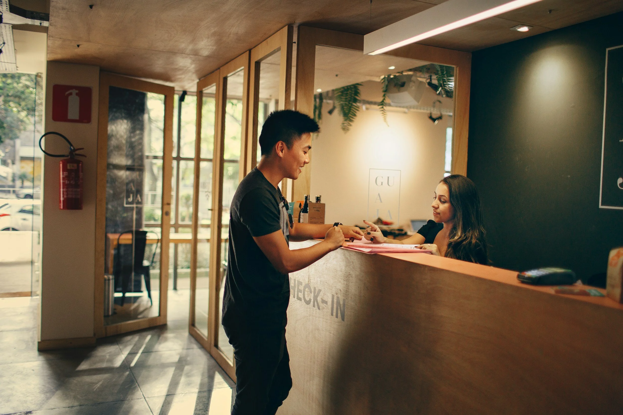 A young man is checking in at a hotel reception desk, handing over a card or forms to a female receptionist in a modern lobby with wooden accents and natural light.