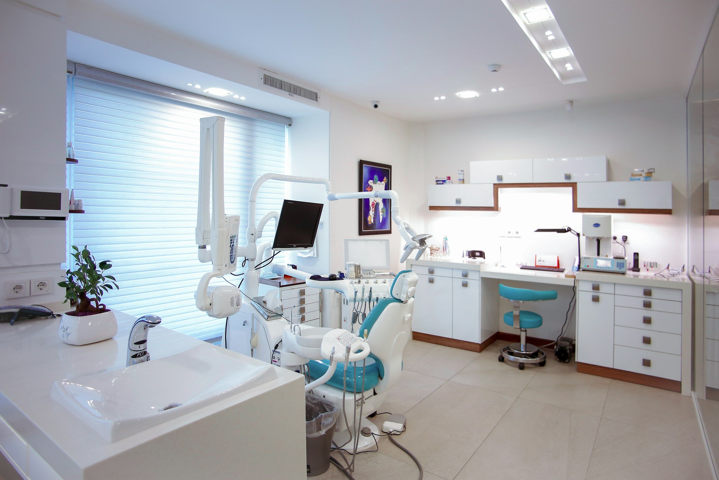 A modern dental office with dental chair, medical equipment, and cabinets, illuminated by bright lighting and a window with blinds.