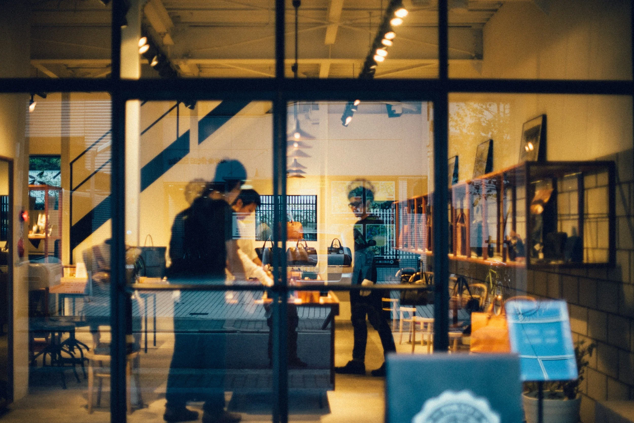 People inside a store or café seen through a large glass window at night, with reflections of interior lighting and objects. Several individuals are visible, engaged in conversation or activities.