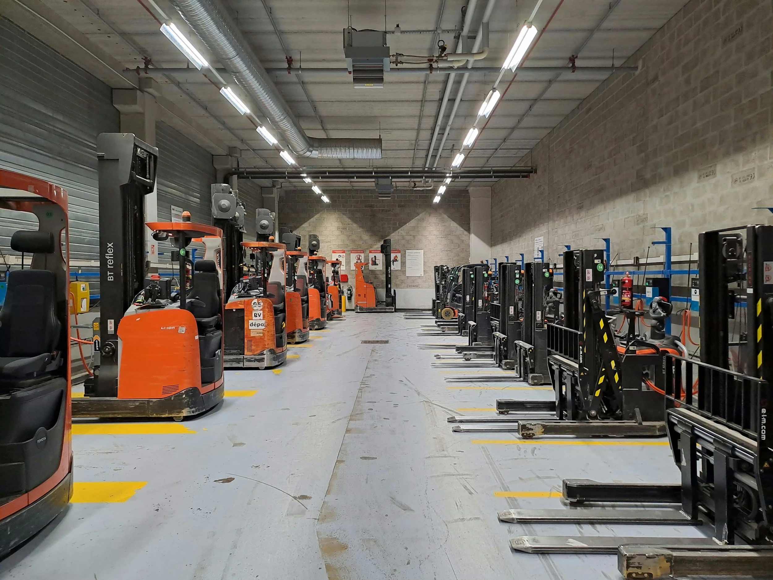 Warehouse with forklifts parked in designated spots, concrete floor with yellow markings, brick walls, and overhead lighting.
