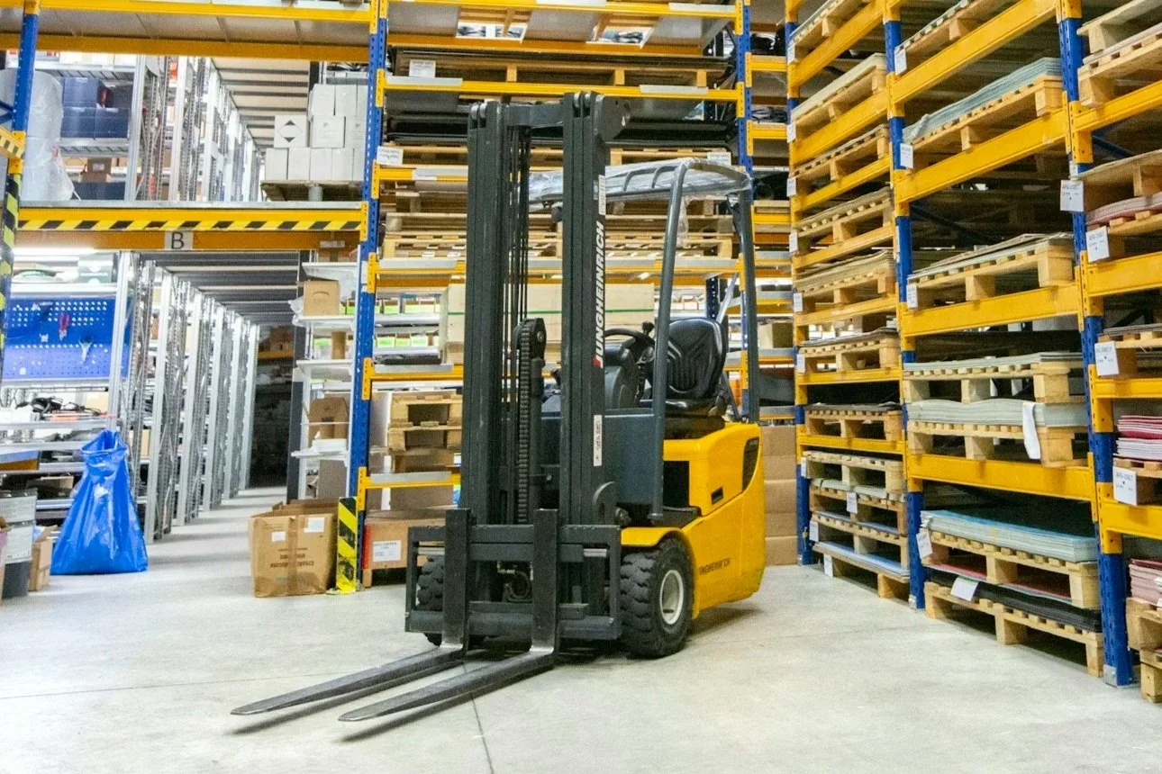 A yellow and black forklift in a warehouse aisle surrounded by shelves stacked with boxes and materials.