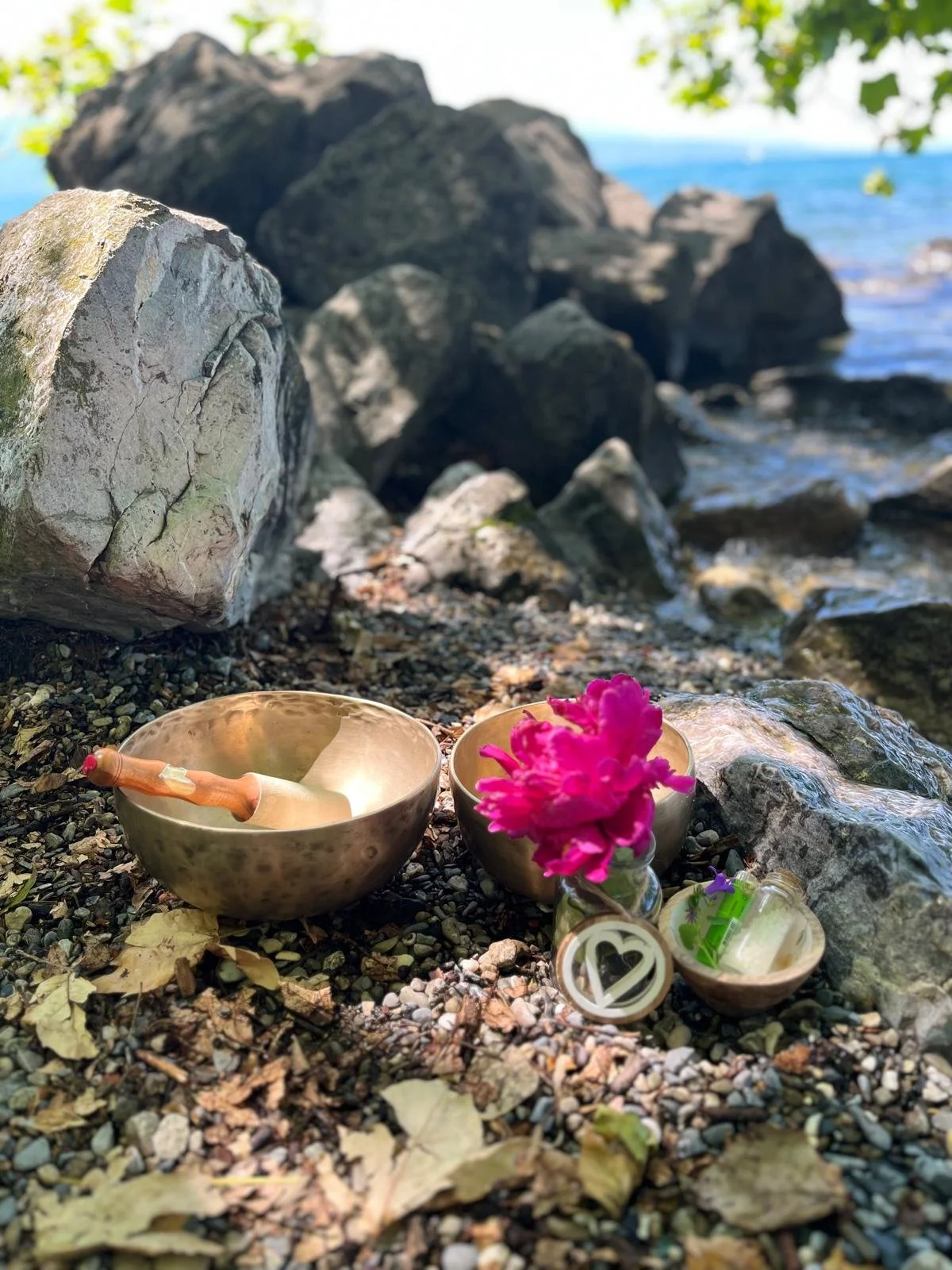 Deux bols de méditation en métal, une petite cuillère en bois, une fleur rose dans un vase en verre, une bougie dans un pot en verre, et plusieurs petits objets autour, posés sur une plage rocailleuse près de l'eau, avec des rochers et des arbres en arrière-plan.