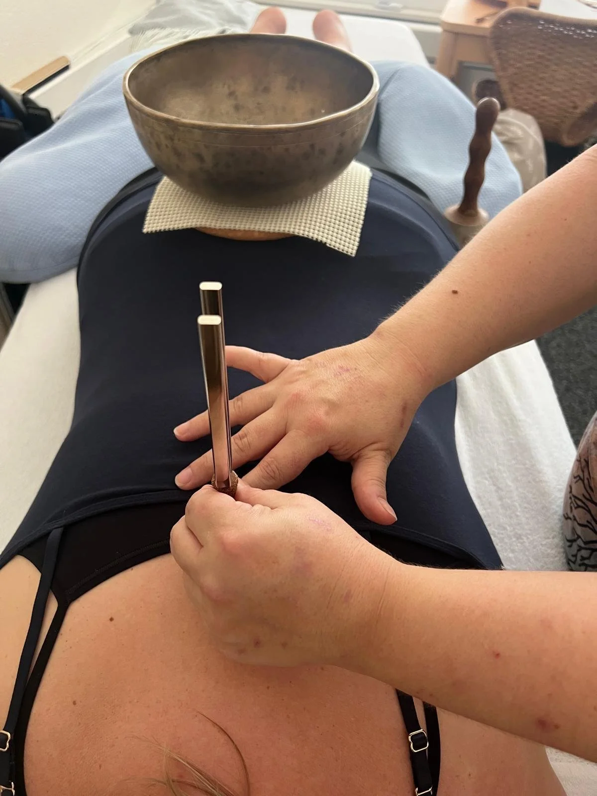 Une personne allongée, recevant une séance de réflexologie ou d'acupuncture avec une baguette en métal, ses mains reposant sur ses jambes, avec un bol en métal placé sur sa cuisse, et un environnement intérieur.