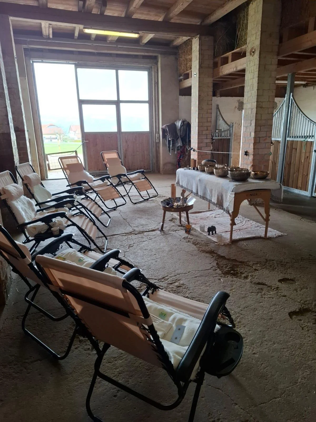 Une pièce en construction avec plusieurs chaises roulantes alignées, une table avec des plats et des objets, murs en brique et grande porte-fenêtre donnant sur un paysage rural.