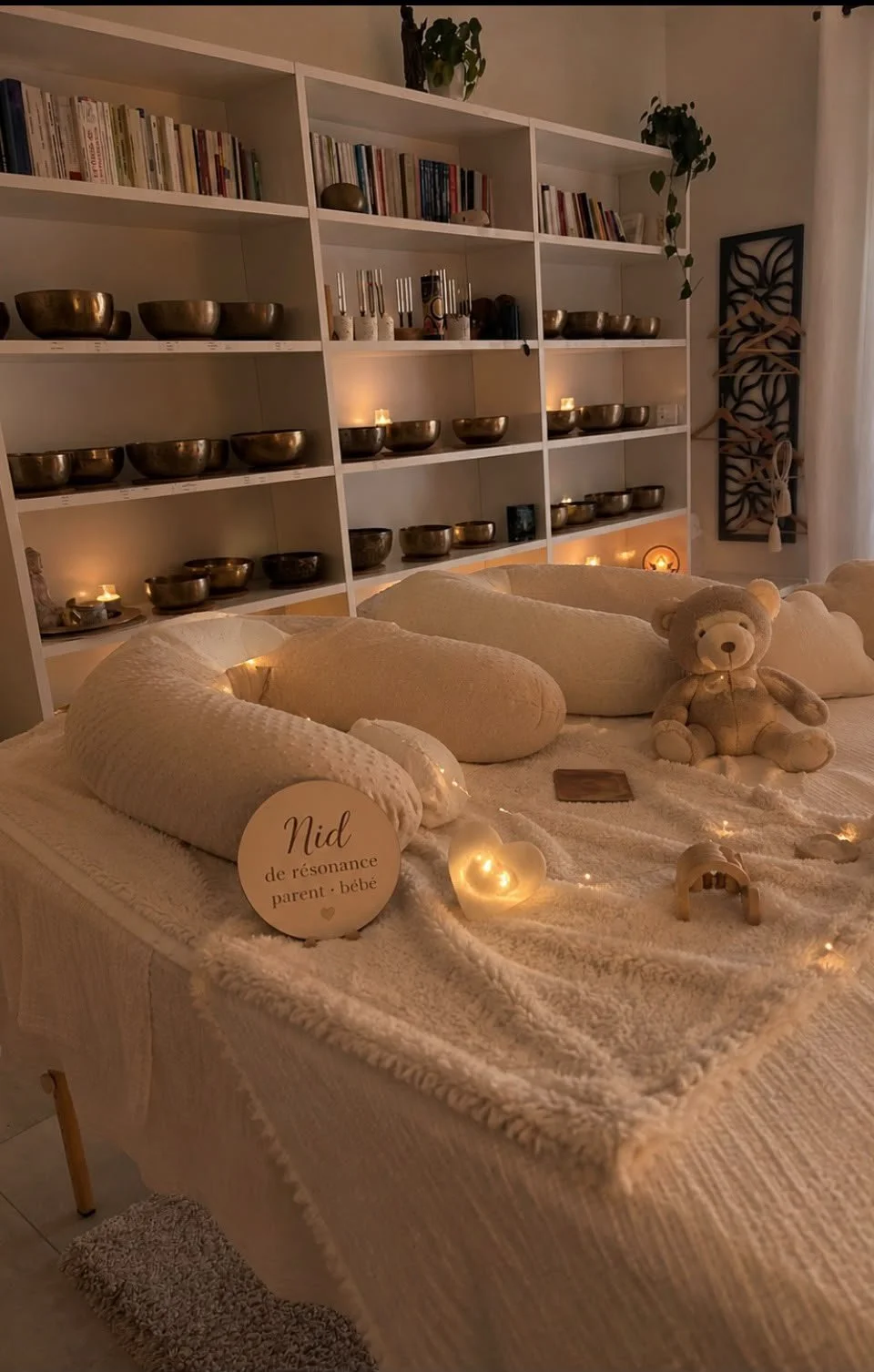 Une pièce chaleureuse avec une étagère contenant des bols en métal, des livres, des bougies, une chaise longue avec des coussins en peluche et un ours en peluche, décorée avec des guirlandes lumineuses et une affiche vintage à l'arrière-plan.