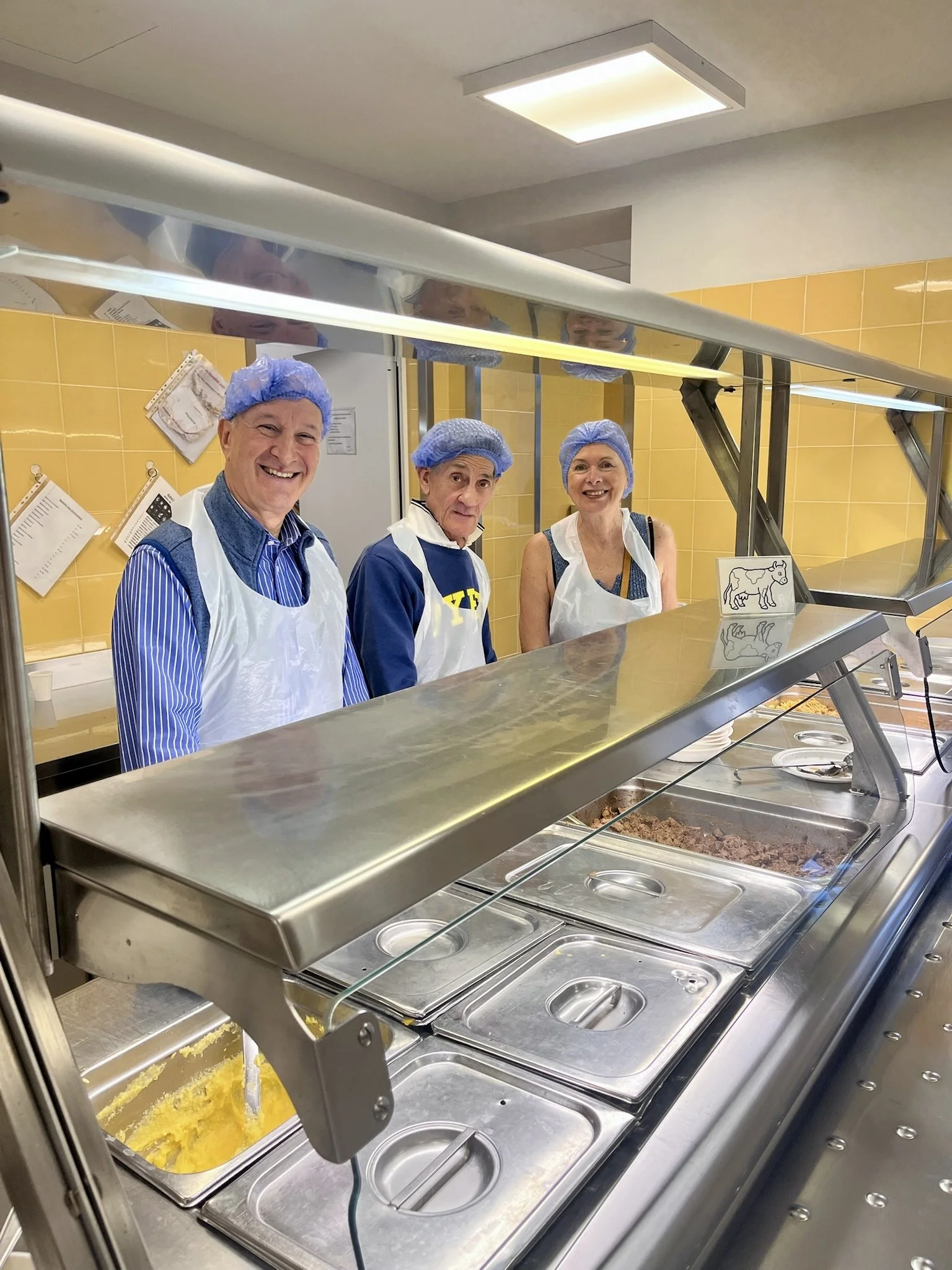 Three people wearing hairnets and aprons stand behind a stainless steel cafeteria serving station with various trays of food, including mashed potatoes and ground meat, in a yellow-tiled room.