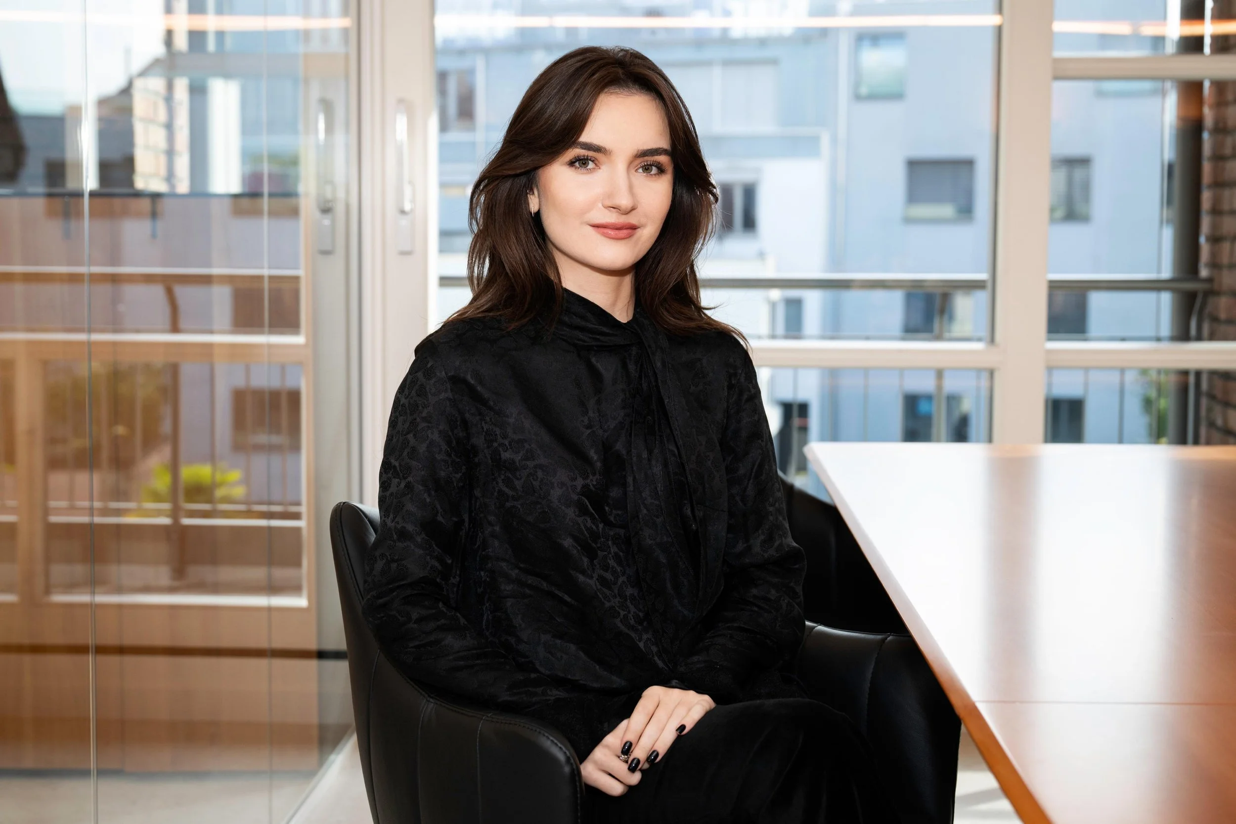 Portrait of a woman with dark brown hair, wearing a black top, standing indoors near a window with city buildings in the background.