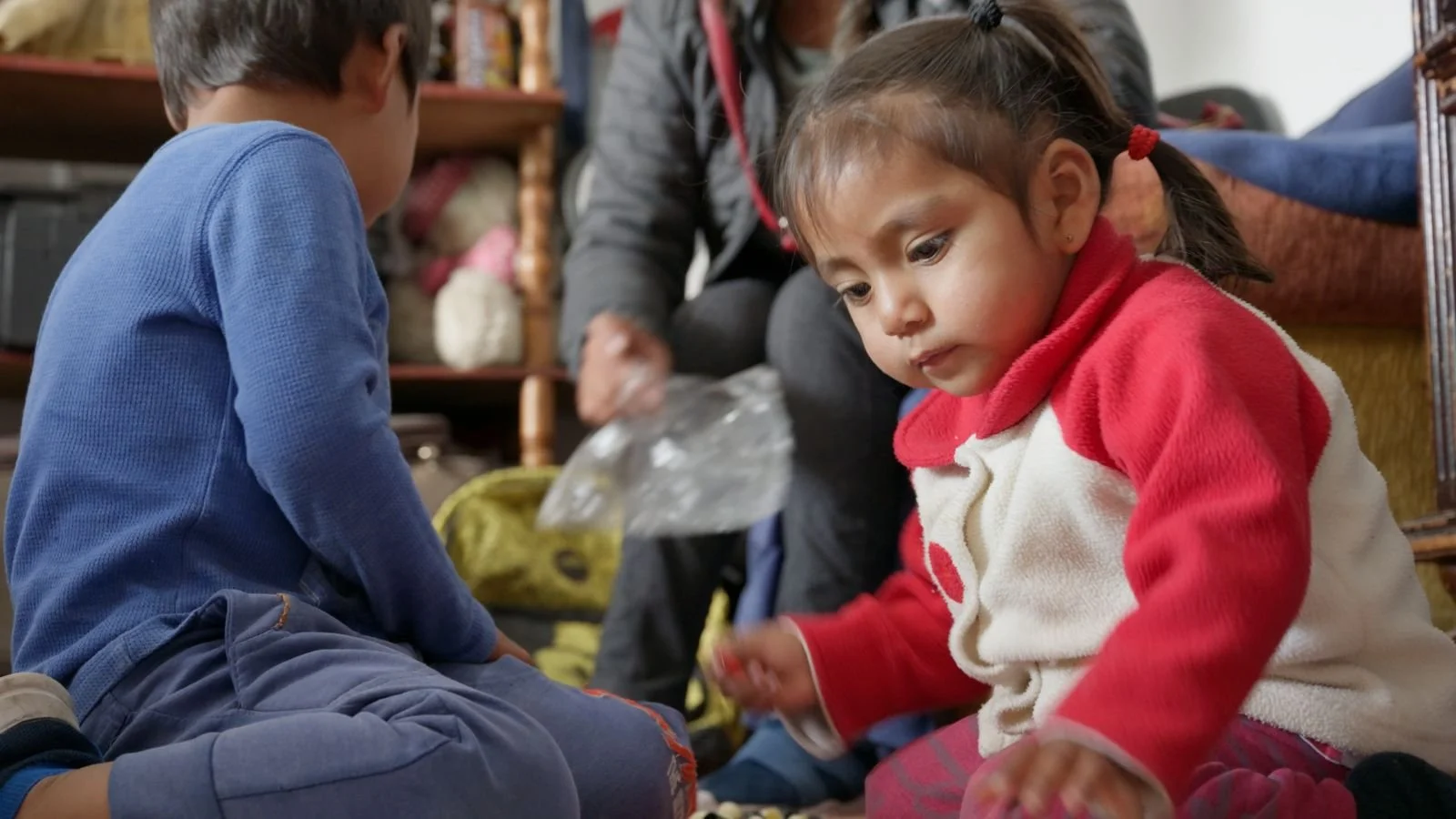 Two children, a boy and a girl, sitting on the floor and playing with small objects while an adult assists them in a living room.