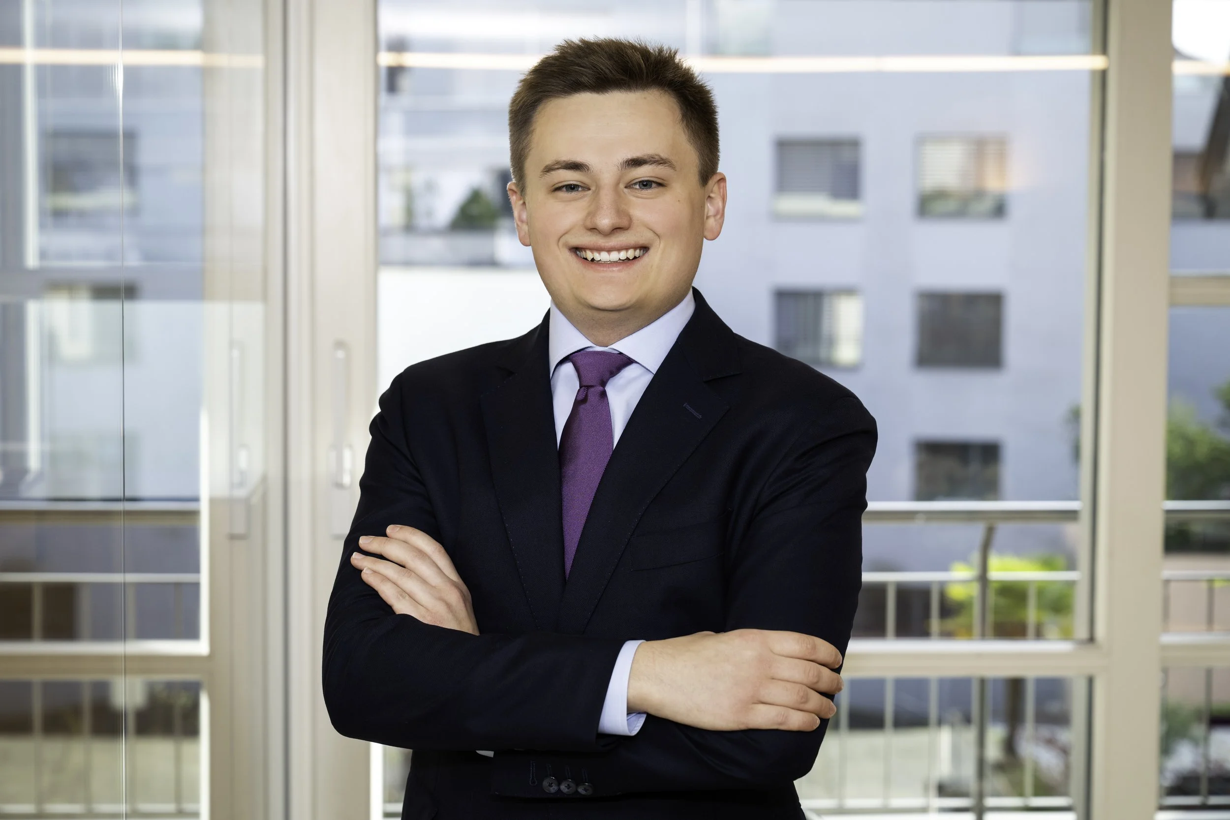 Professional young man in suit smiling in an office with large windows.