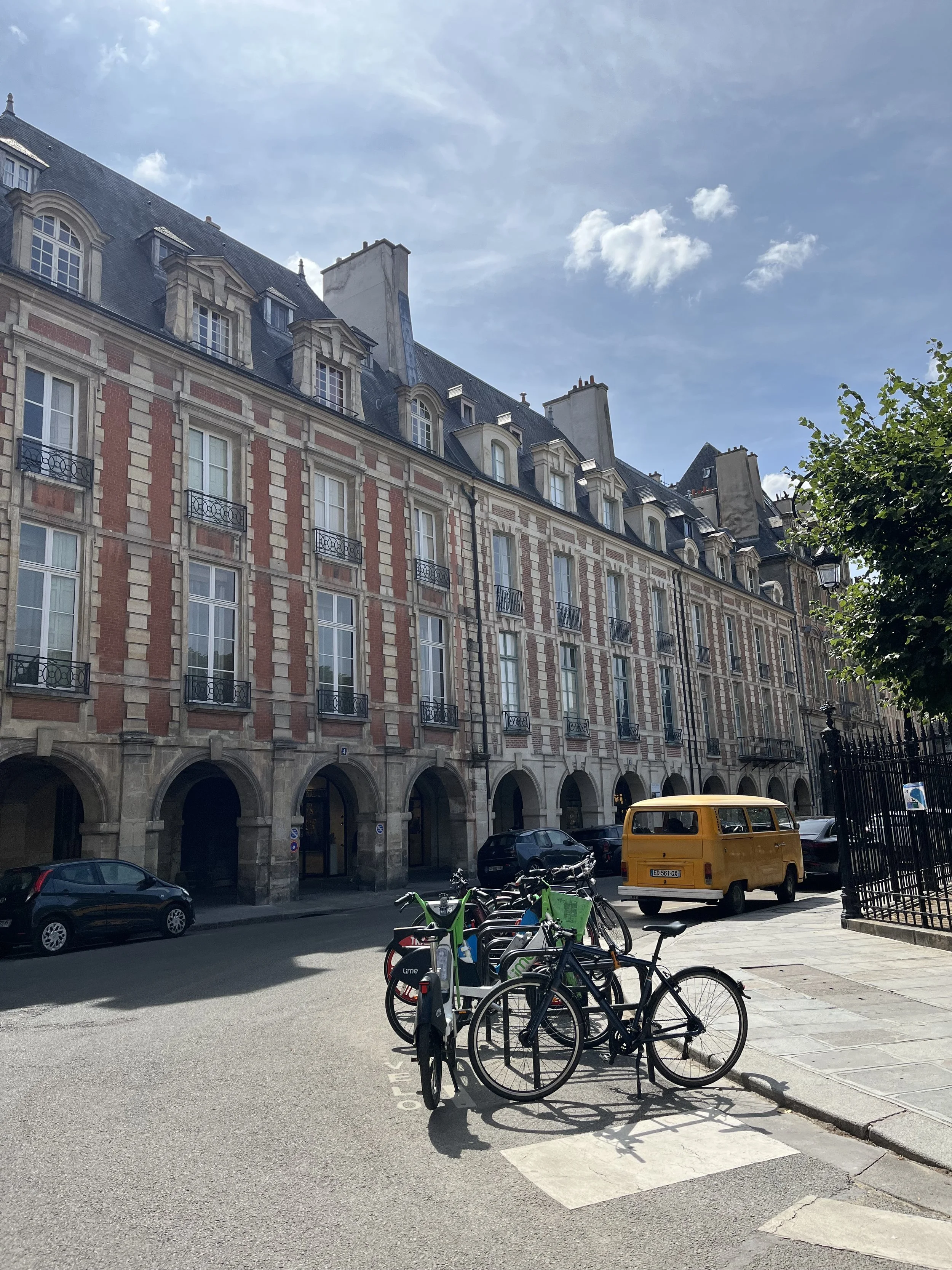 Rue animée avec des vélos stationnés, une voiture jaune et plusieurs voitures noires, immeuble haussmannien avec des fenêtres et balcons en fer forgé, ciel bleu avec quelques nuages.