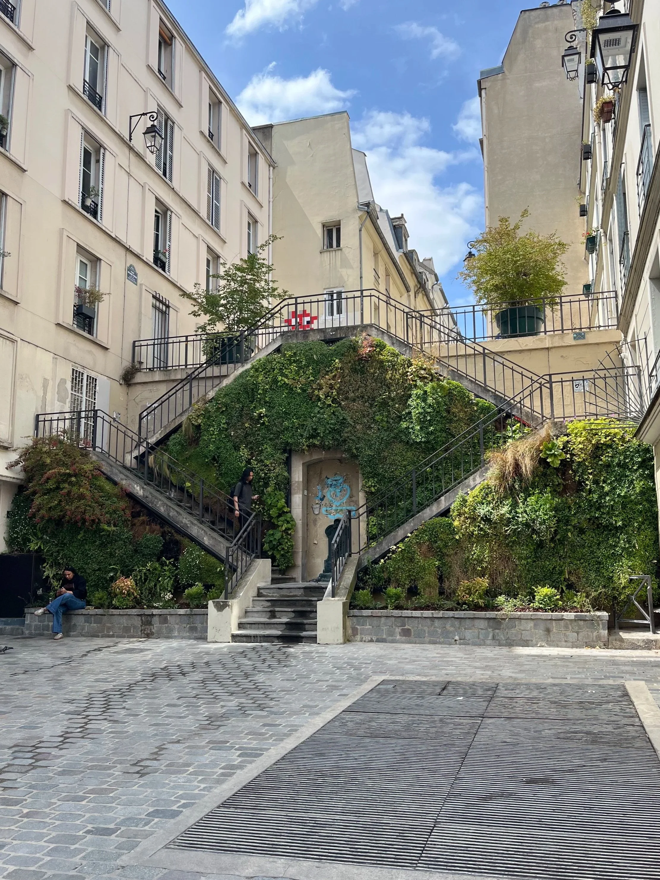 Une place pavée au centre, avec une montée d'escalier menant à un espace vert rempli de plantes et de buissons. Deux personnes sont visibles, une assise sur un mur et une d'une marche, dans un environnement urbain avec des bâtiments anciens et des lampadaires en métal.