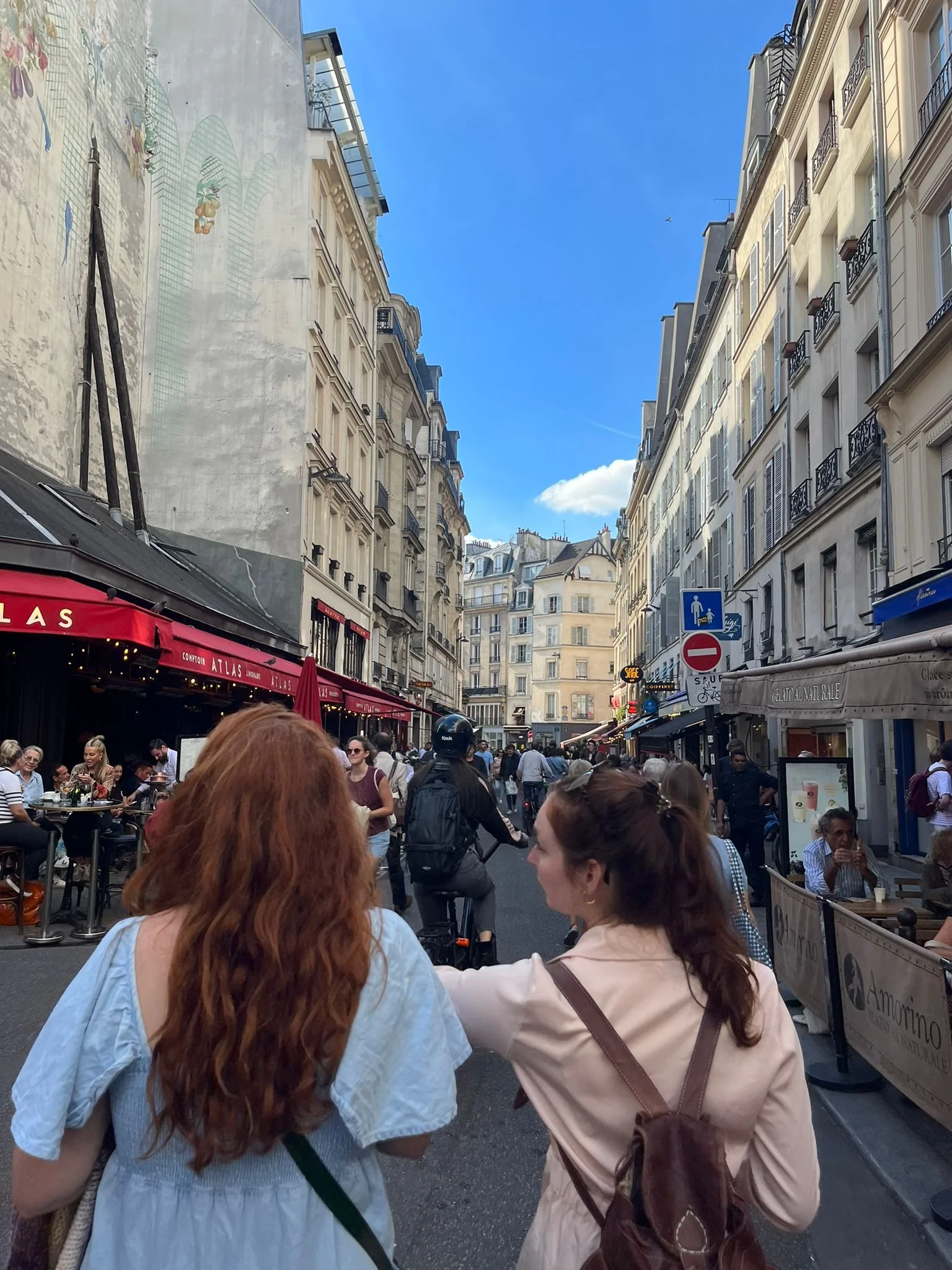 Rue animée à Paris avec des bâtiments haussmanniens, des cafés avec terrasses, des panneaux de signalisation, et de nombreuses personnes, notamment deux femmes main dans la main au premier plan, sous un ciel bleu avec quelques nuages.