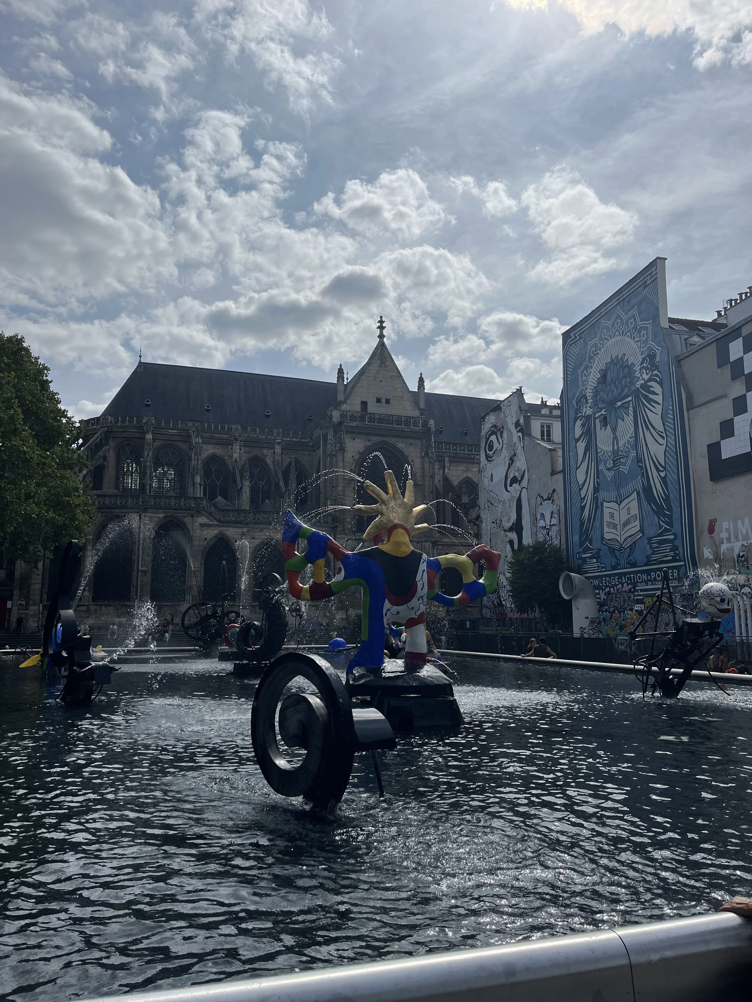 Une fontaine dans une place publique avec des sculptures colorées, un bâtiment historique en arrière-plan, des affiches géantes avec des illustrations en bleu et blanc, et un ciel partiellement nuageux.
