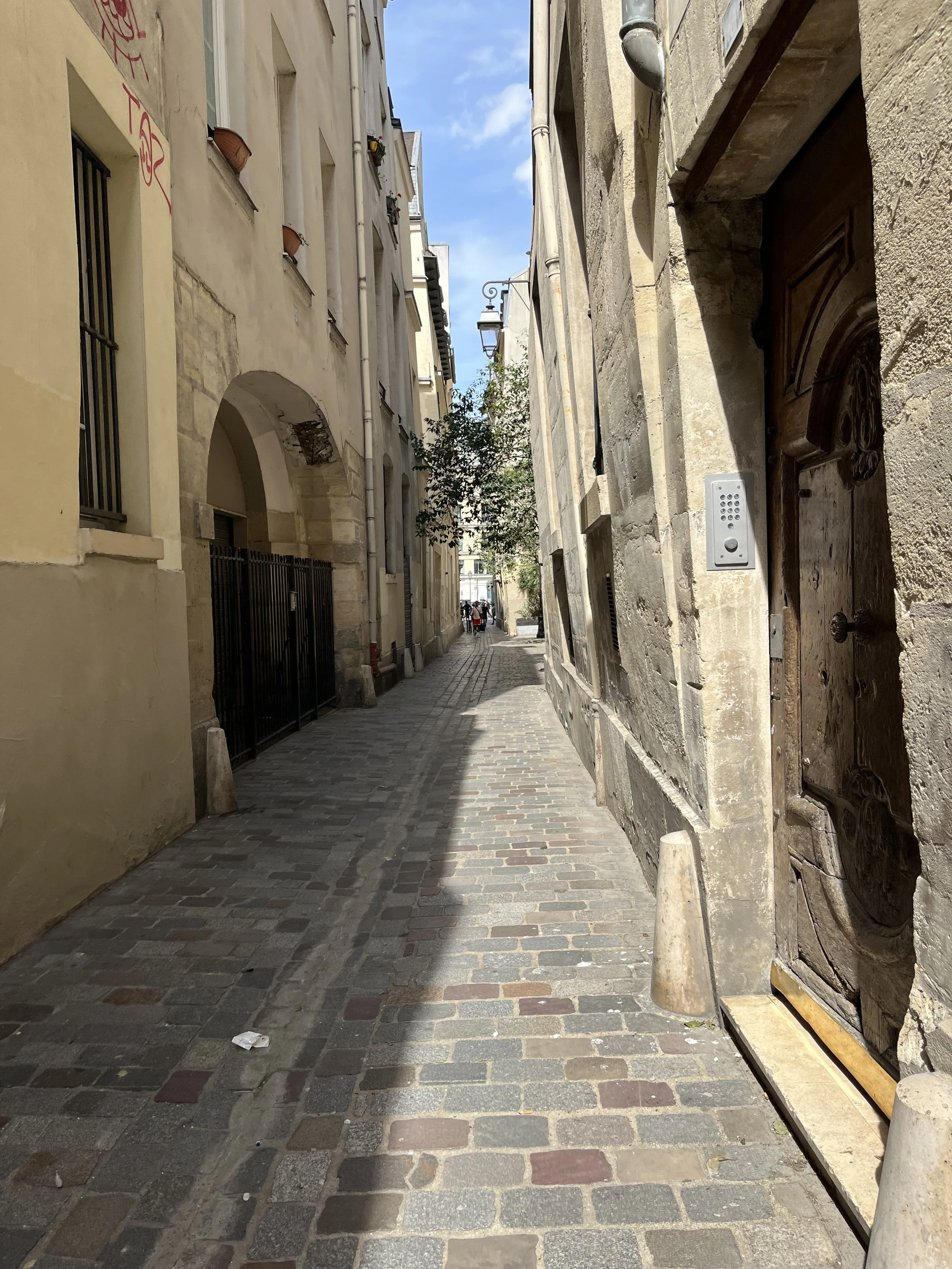 Une ruelle pavée étroite bordée de bâtiments anciens en pierre et en stucco, avec un lampadaire suspendu au-dessus de la rue. Le ciel est bleu avec quelques nuages, et quelques personnes sont visibles au fond de la rue.