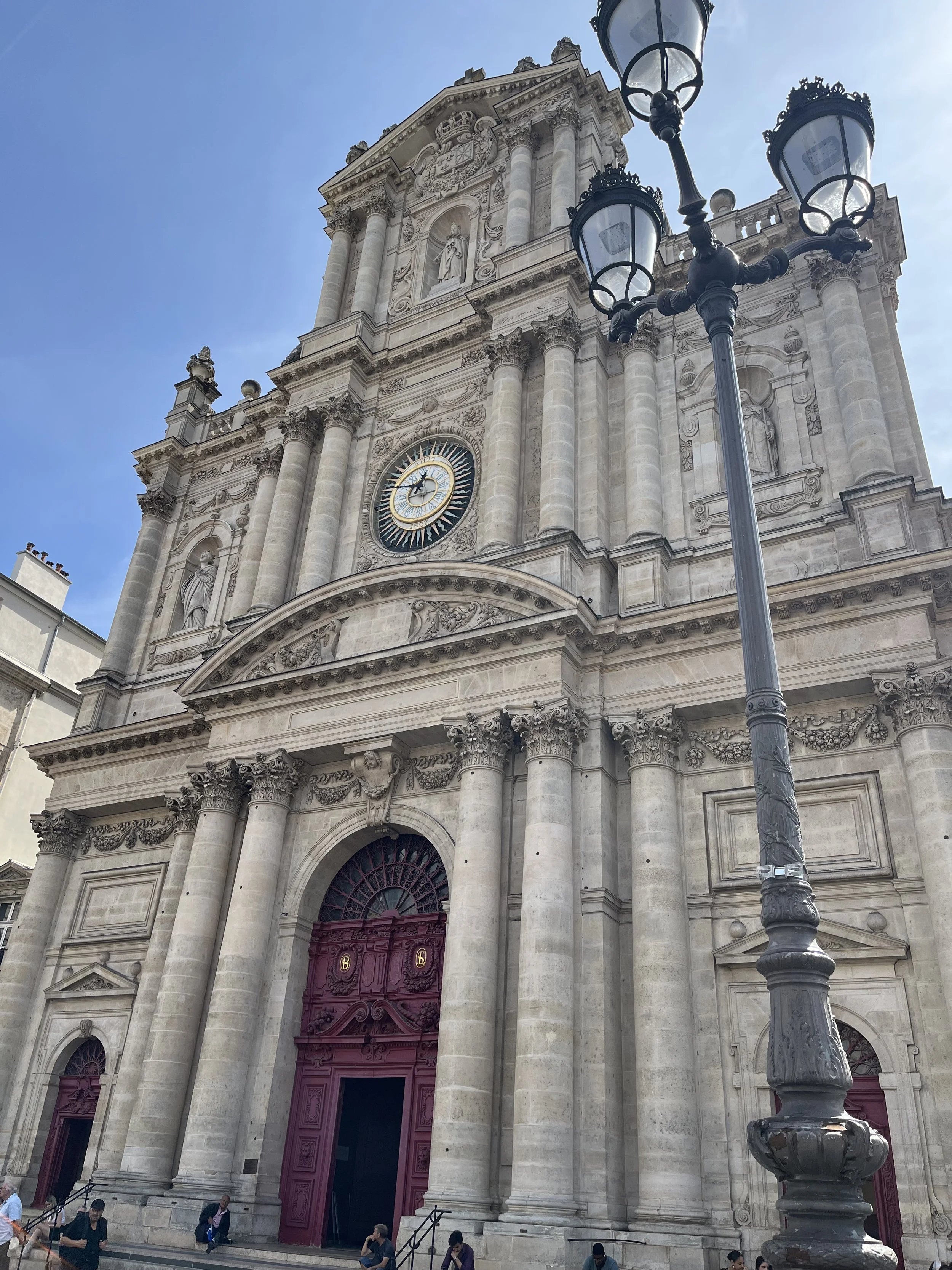 Faculté de style classique, façade imposante en pierre avec colonnes et statues, portail en bois violet, horloge au sommet, lampadaire en avant.