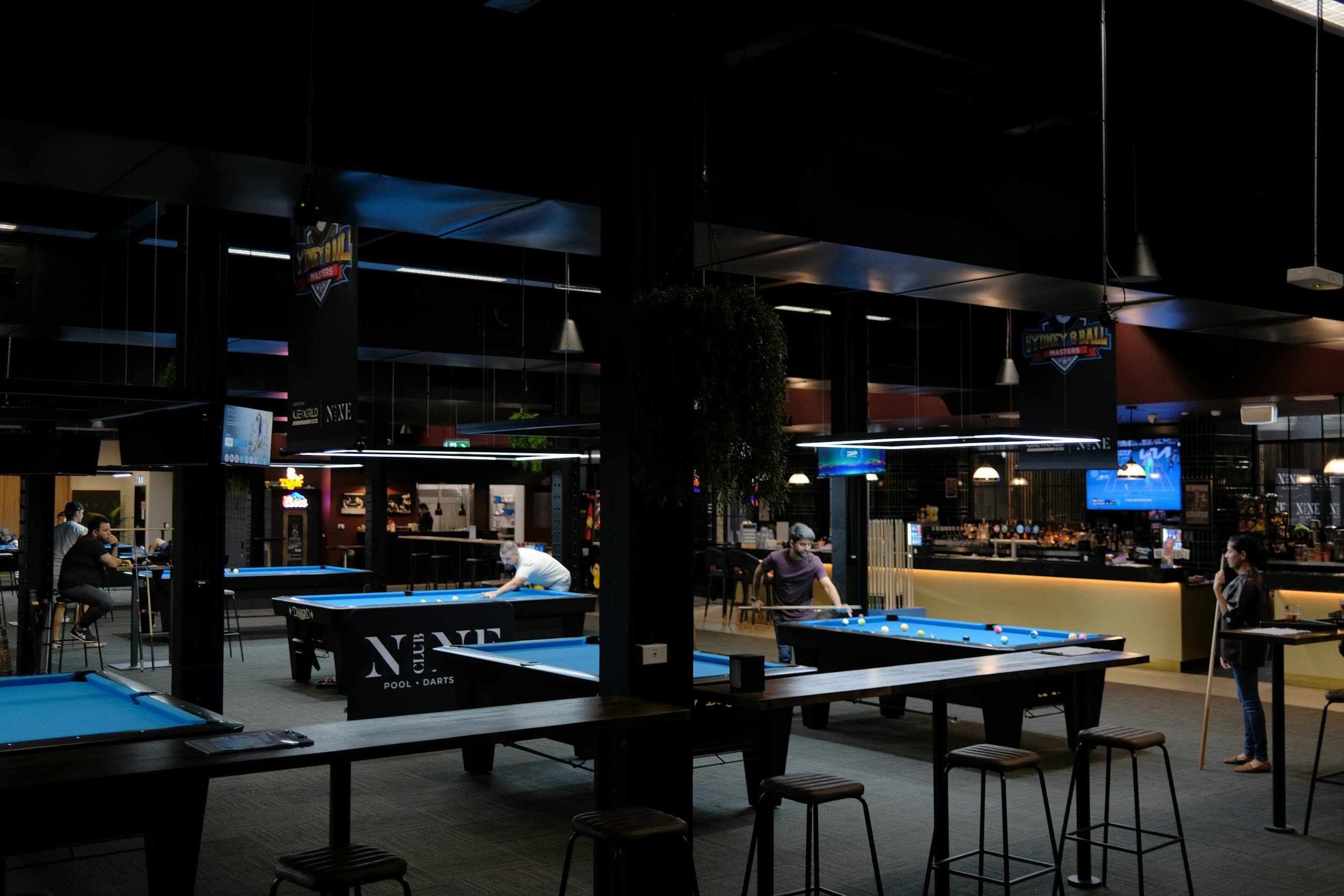 Indoor billiards hall with multiple pool tables, people playing, and dim lighting with signage on the walls.