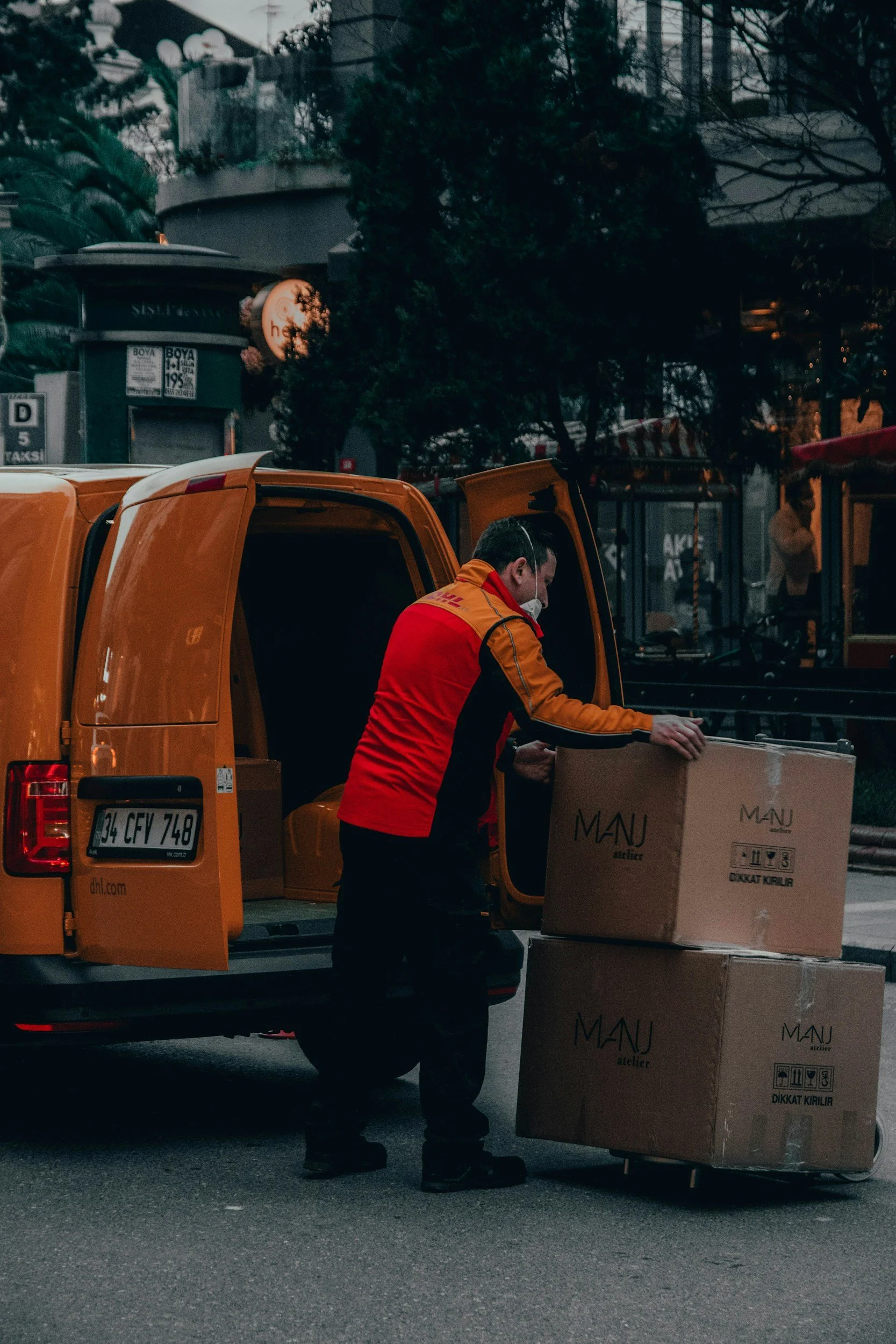 A delivery person wearing a red and yellow uniform and a face mask is loading large cardboard boxes into the back of an orange DHL delivery van parked on a city street. The scene is set during the evening with trees and city buildings visible in the background.