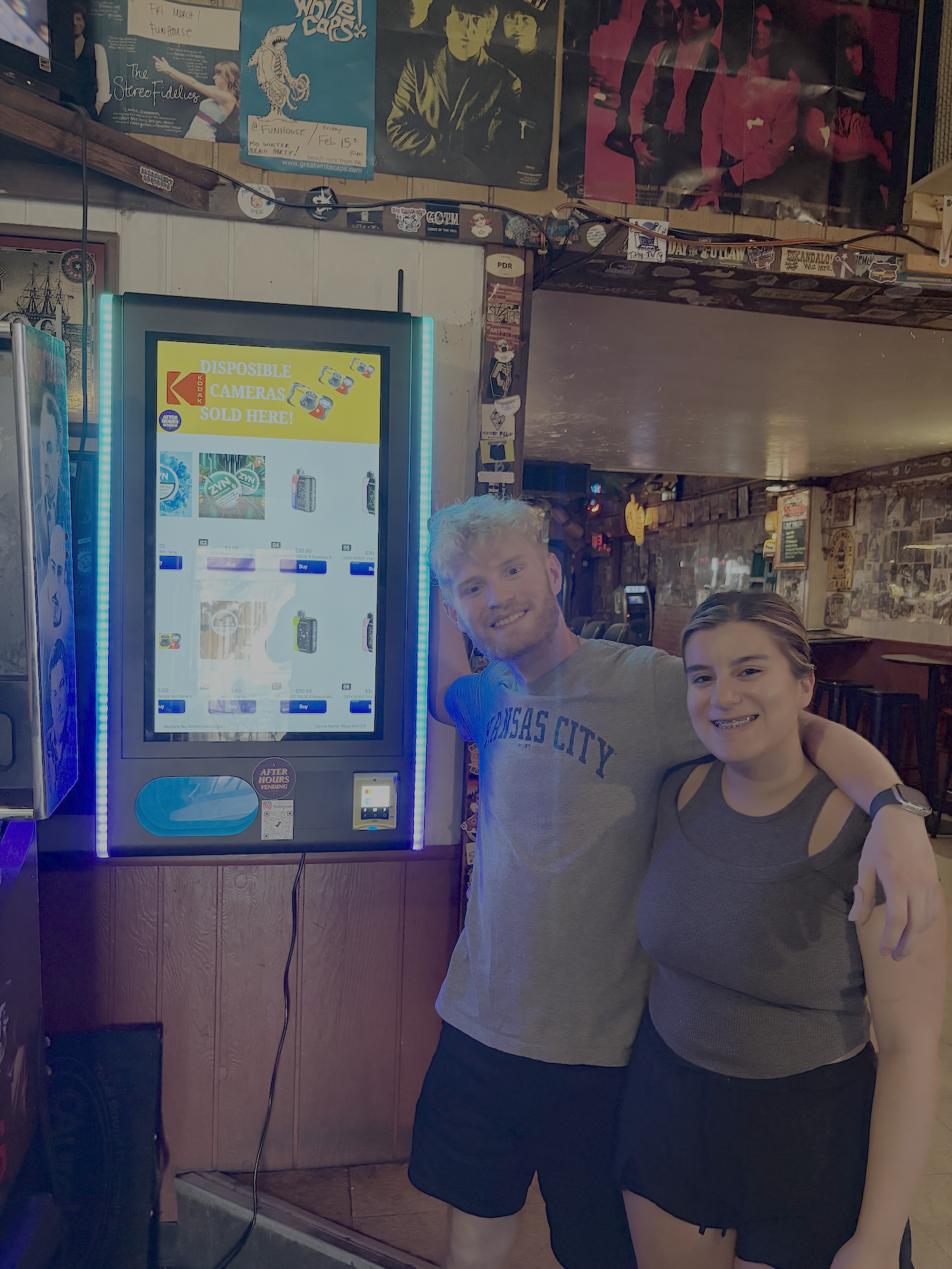 Two smiling young adults, one male and one female, standing together in front of a vending machine inside a bar or restaurant. The background features posters on the wall and a wooden interior.
