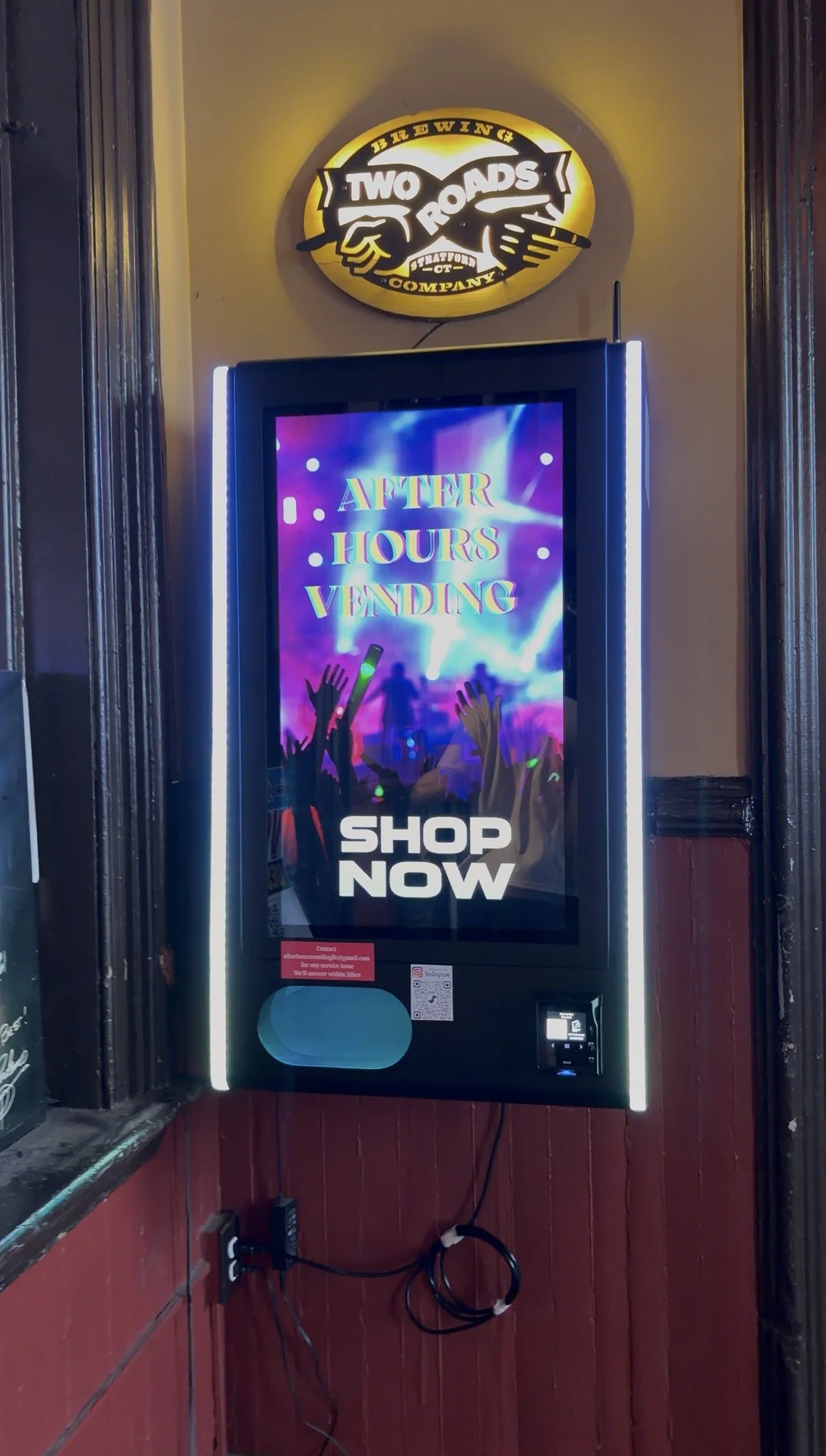 Digital vending kiosk with a colorful screen displaying 'After Hours Vending' and 'Shop Now', positioned against a wall with a sign above reading 'Two Roads Brewing Company' featuring a logo of a yellow oval with a black fist and barbell.