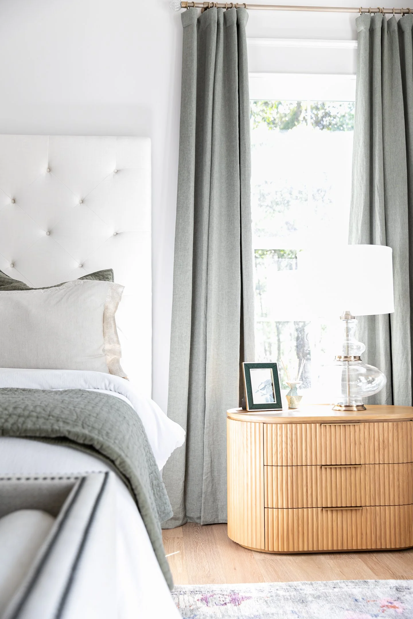 A bedroom corner with a white tufted headboard, a nightstand with a framed photo, a glass lamp, and a window with light gray curtains.