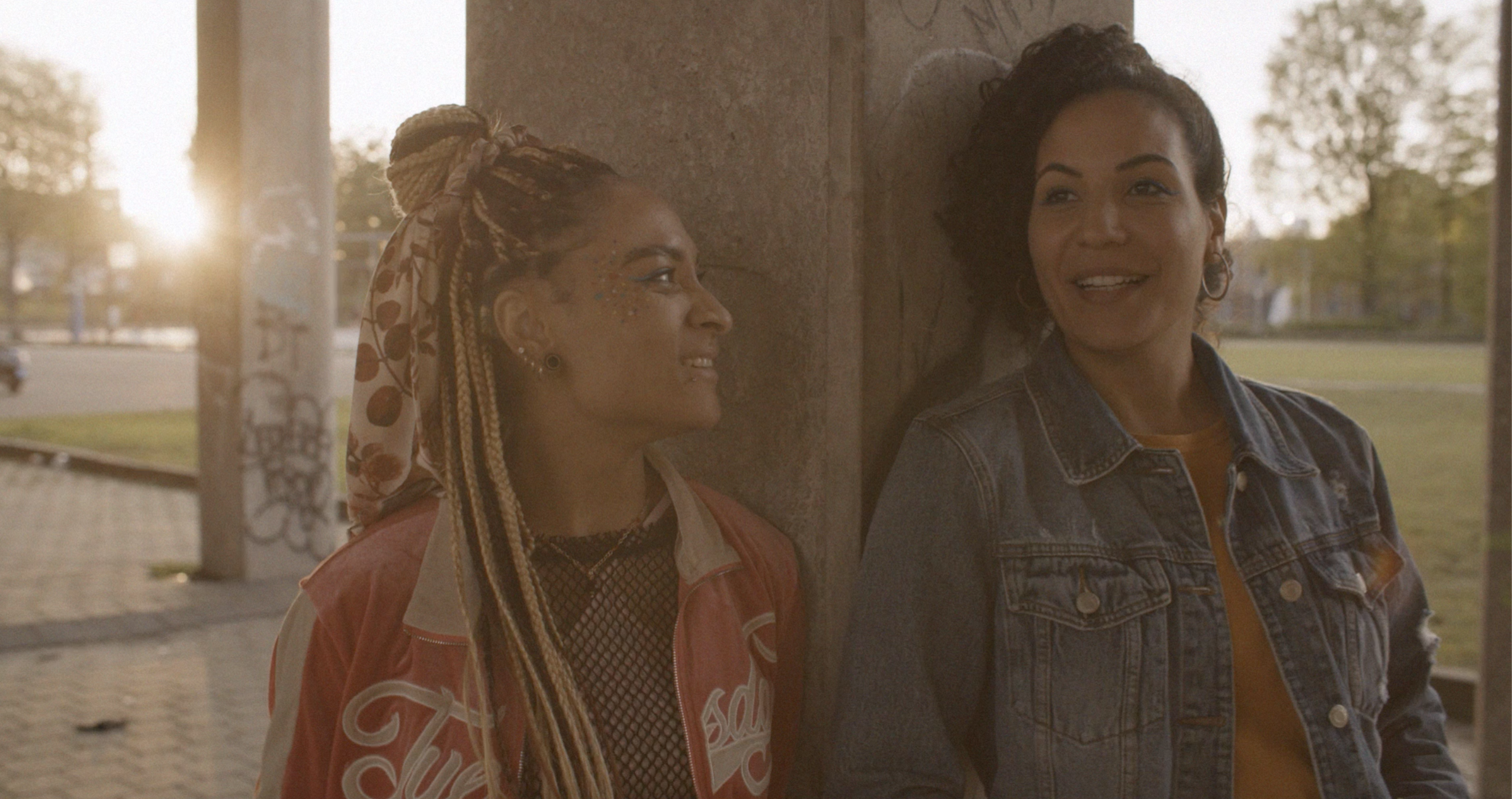 Two women with different hairstyles and skin tones leaning against a wall outdoors, smiling and talking, with sunlight in the background.